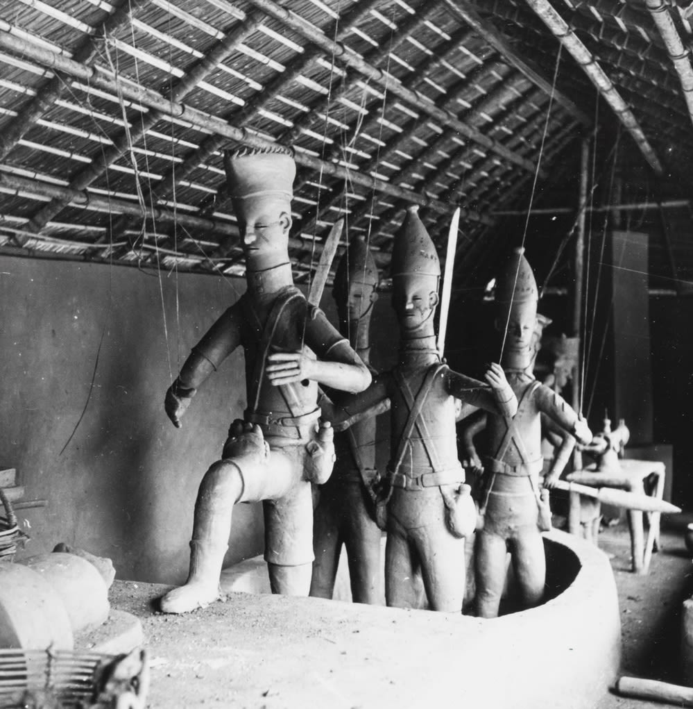 A group of clay figures standing in a boat inside a thatched-roof structure; several figures have swords in their backpacks behind.