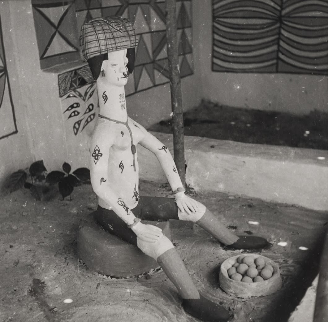 A seated clay figure wearing a patterned head covering rests on a low base, with a shallow bowl of round objects placed on the ground nearby.
