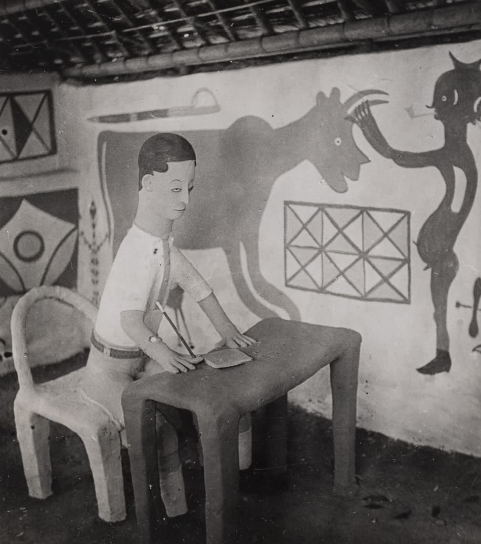 A clay figure seated at a table holding a writing implement, positioned in front of a painted wall featuring animals and geometric motifs.