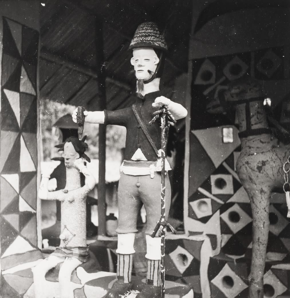 Standing clay figure holding a snake in one hand and a knife in the other, positioned inside a shrine with painted geometric walls; a smaller figure stands beside.