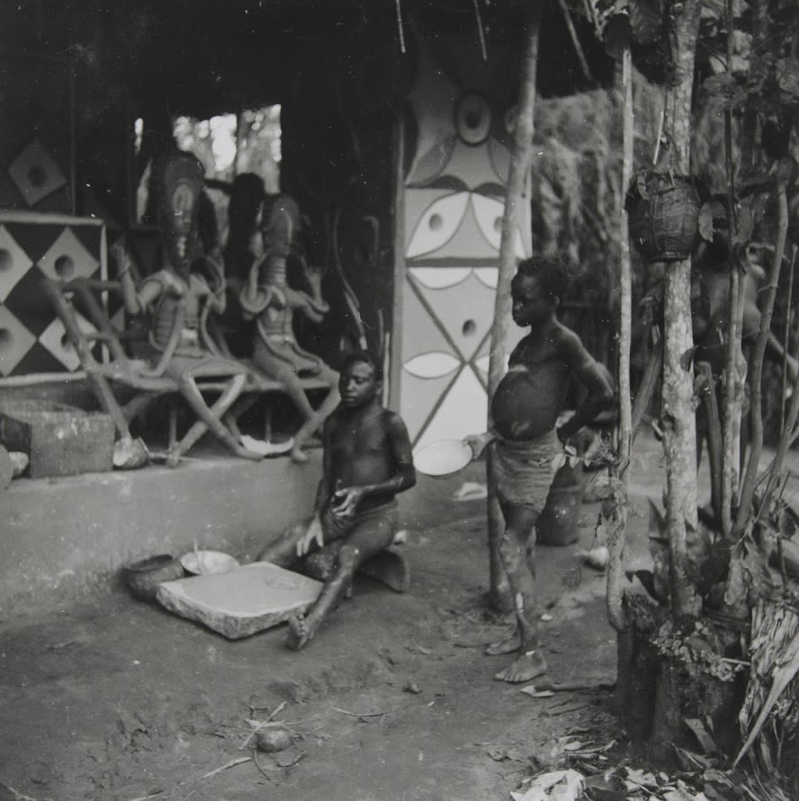 Two people standing and kneeling outside a painted building; sculpted figures sit on a raised platform behind them, with patterned columns and walls visible.