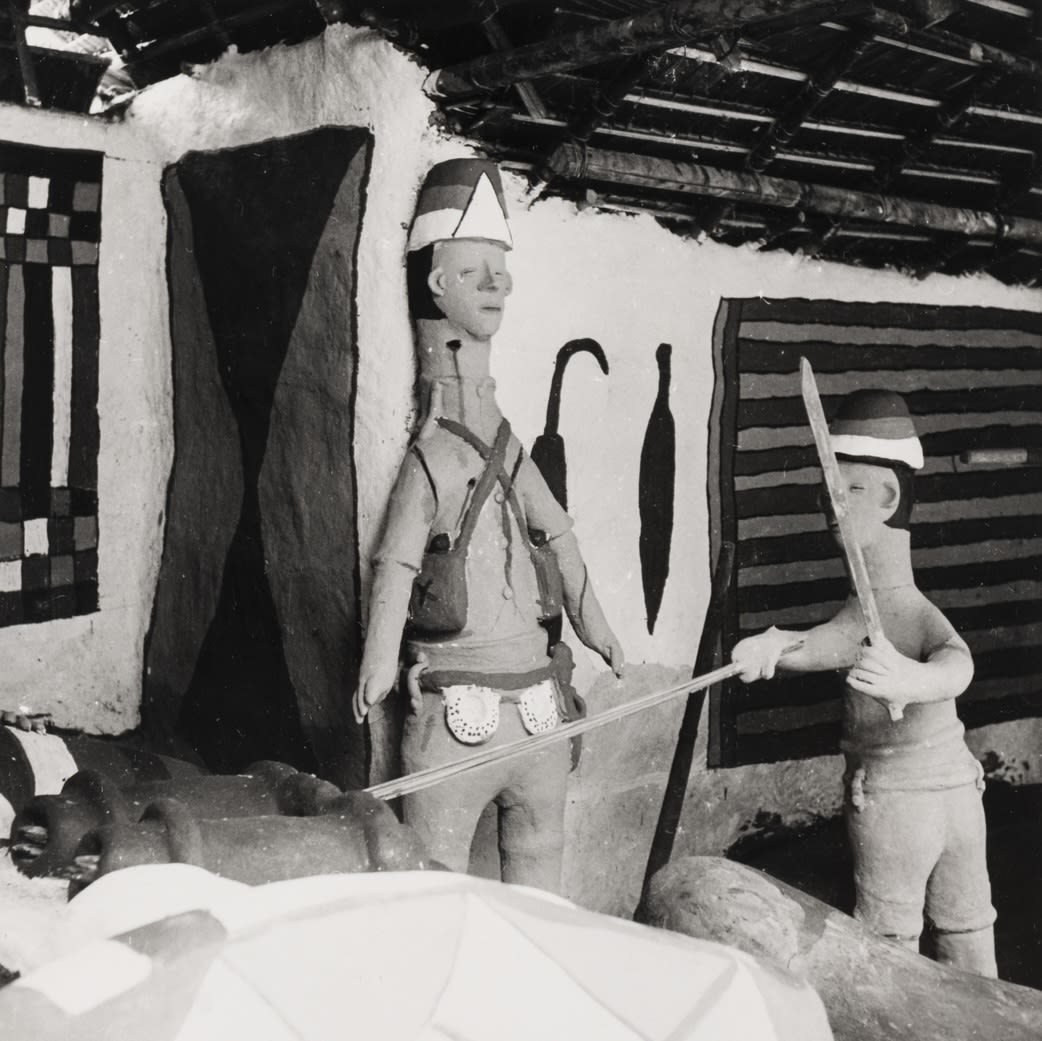 Two clay figures face one another indoors; one holds a long blade while the other holds a shorter object, set against painted walls beneath a thatched roof.