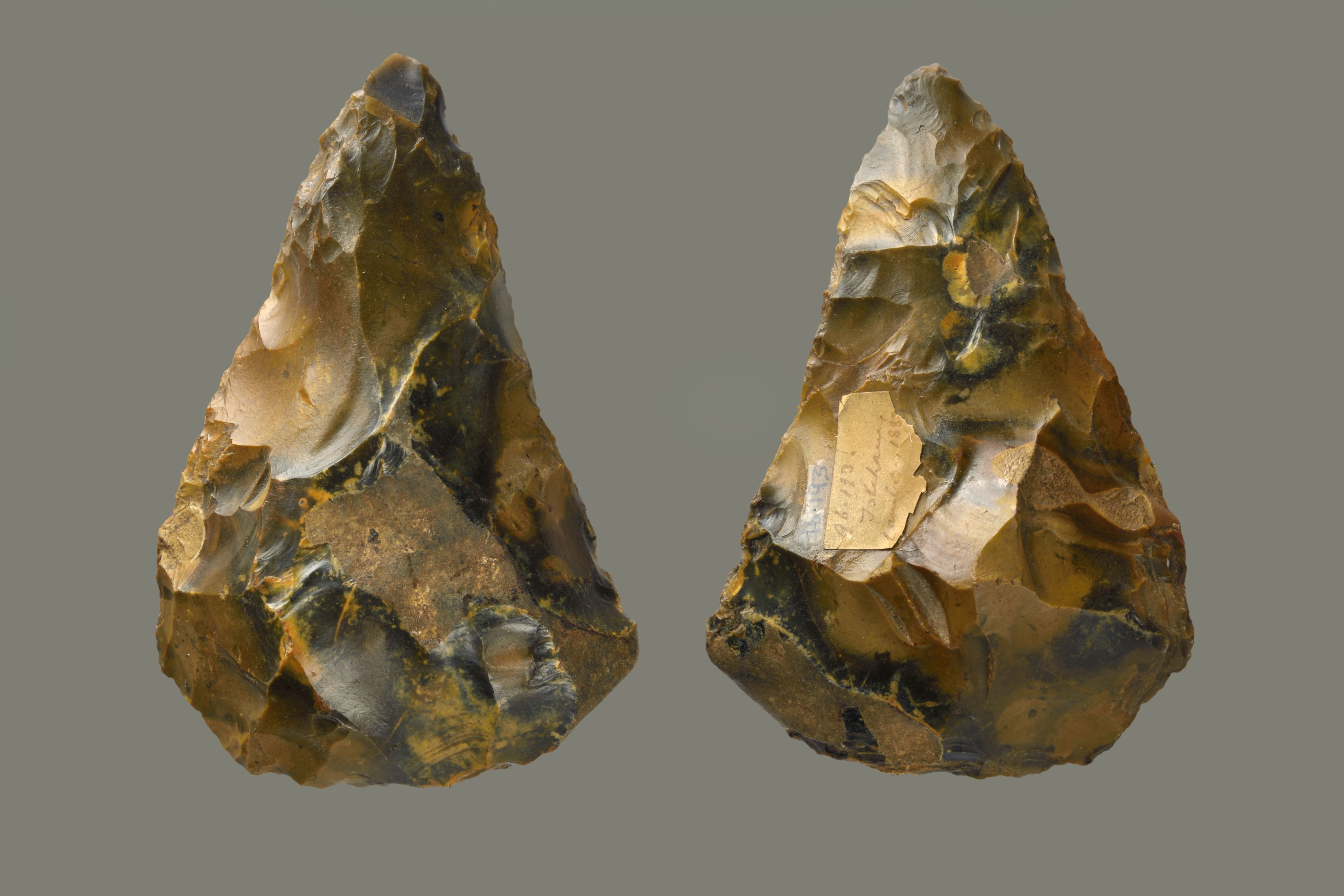 A double-view of a regular sized flint, showing front and back. Triangular in shape, coming to a rounded point at the top. The flint has been chipped away all over leaving marks, and the stone is a rich golden colour, with streaks of dark brown/black through it.