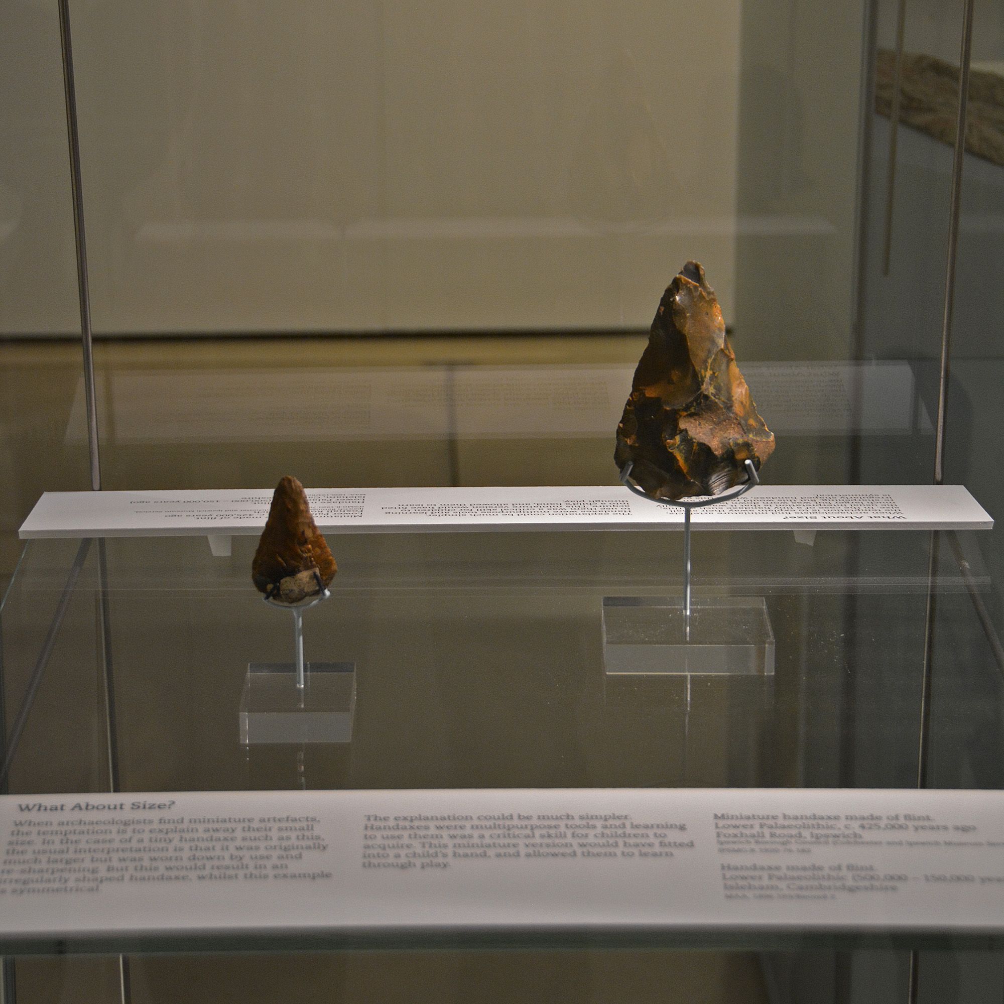 Two flint handaxes arranged in an exhibition case, with perspex bases. One is about double the size of another, smaller one. An out-of-focus text panel is on view below, with the same text as on this slide.