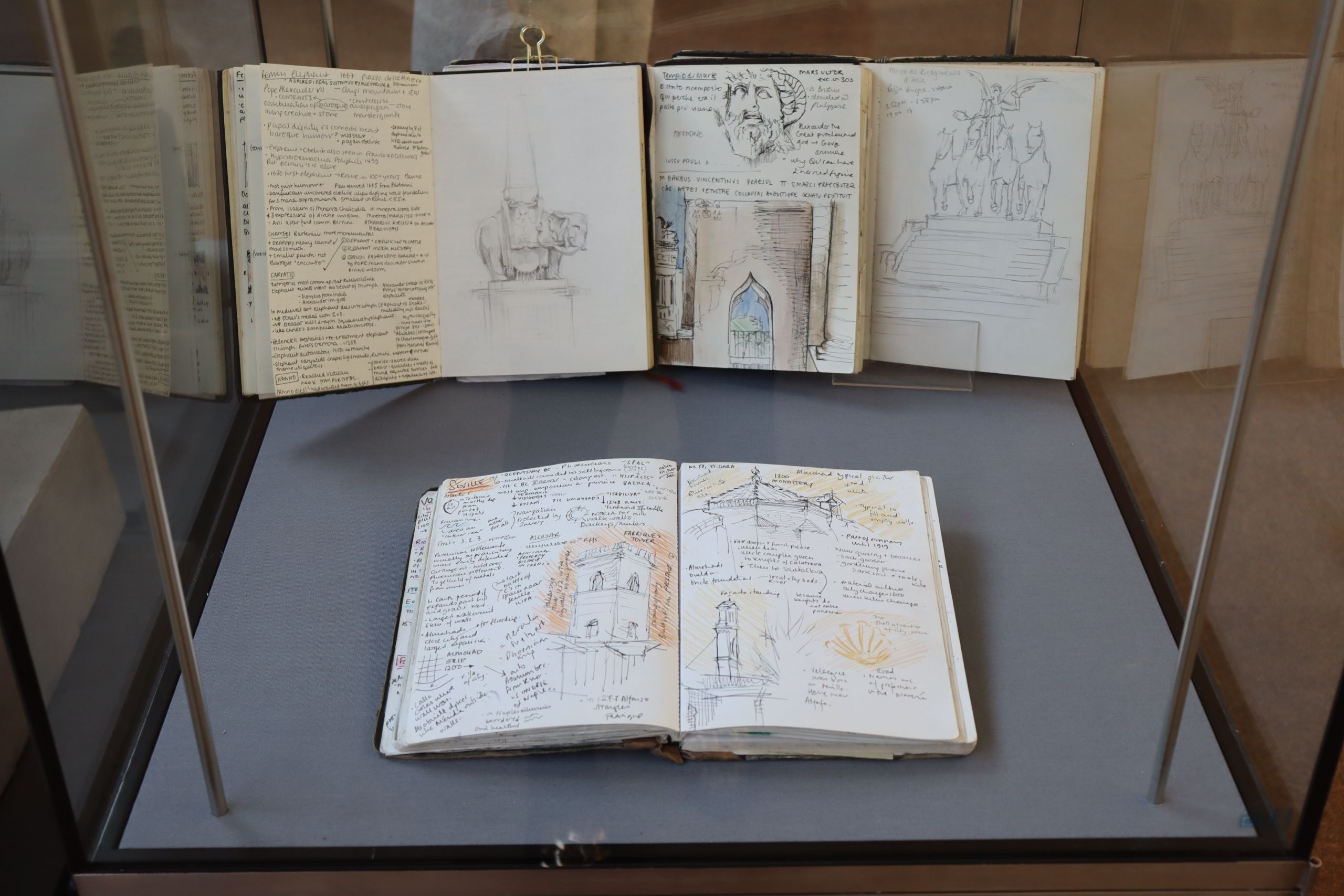 A photograph of a display case with sketchbooks laid open revealing drawings of buildings and statues alongside lots of handwritten notes