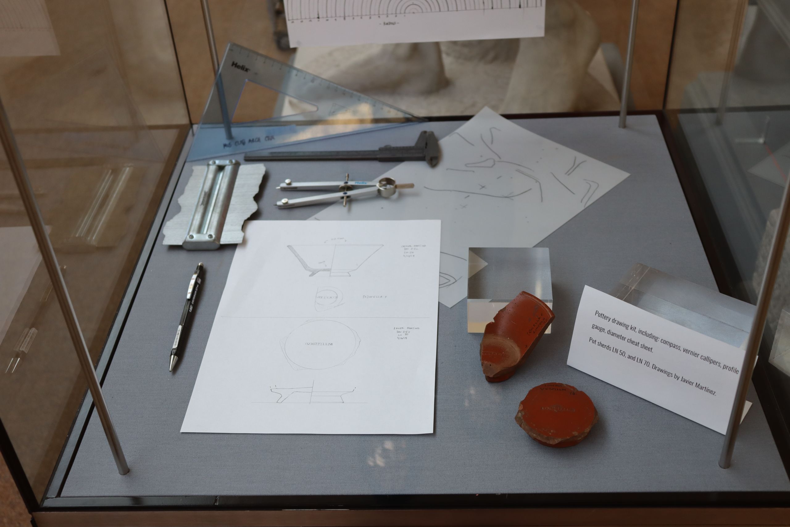 A photograph of a display case, inside is a pottery drawing it, to small broken pieces of Samian ware pottery, drawings of the pottery and a label.