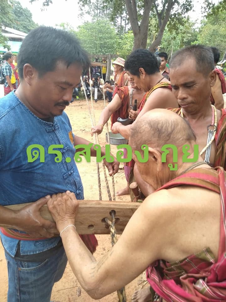 Three men stand holding a block of wood with three holes drilled into the middle. The three strands of buffalo hide leather are threaded through each of the holes and braided together on the other side of the wood. In the background are three more men standing behind the block of wood, helping to pass the long threads of leather through the wooden block.