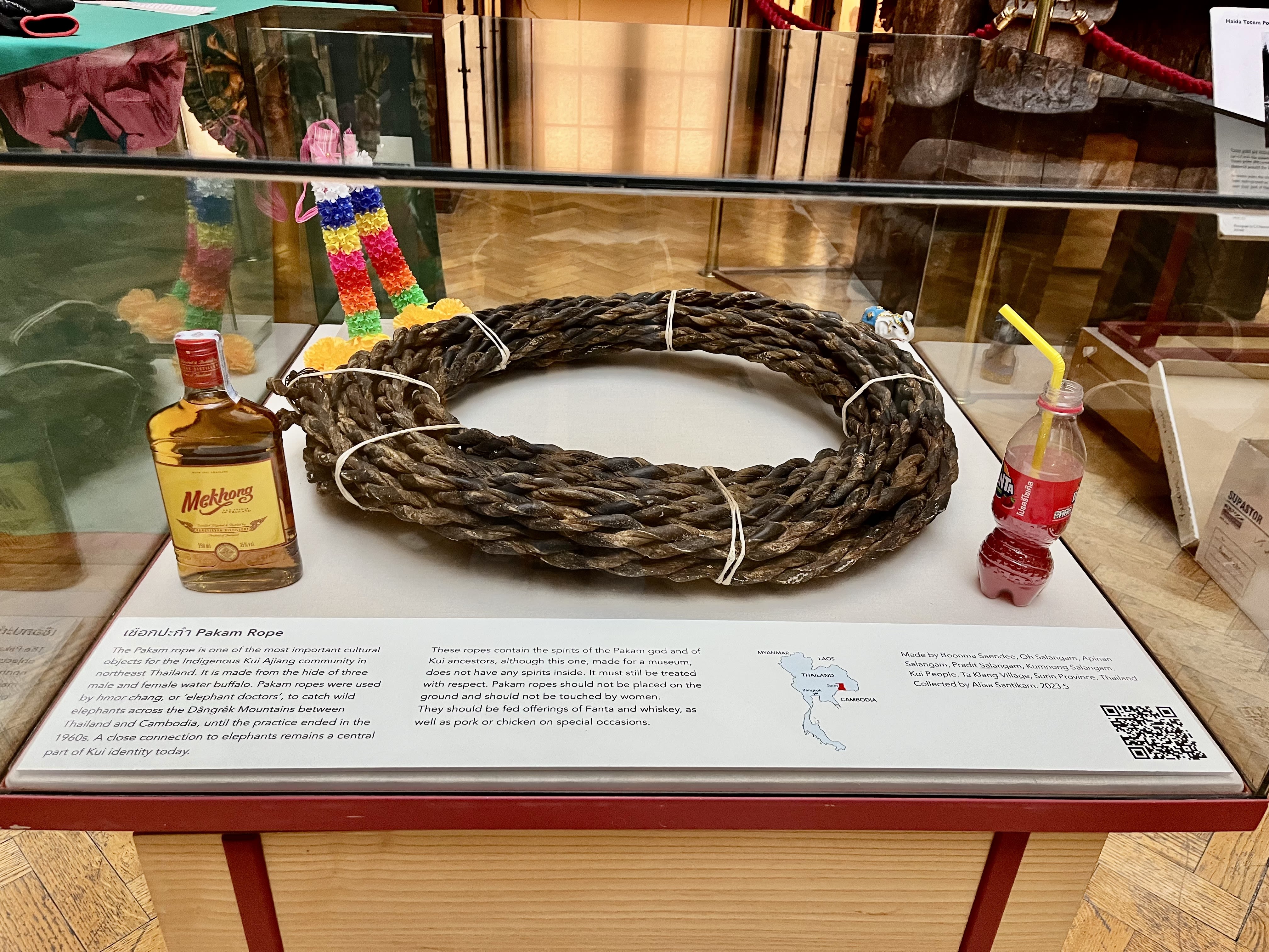 The dark brown woven Pakam rope sits coiled at the centre of the glass display case. On the back left is a plastic replica of a floral garland, with white, blue, yellow, red, orange, and green stripes. In the back right is a small statue of an elephant in white, with its trunk raised and a blue piece of fabric draped on its back. In the front right is an opened bottle of red Fanta, partially filled, with a yellow straw sticking out of it, and in the front left is a bottle of Mekhong whiskey.