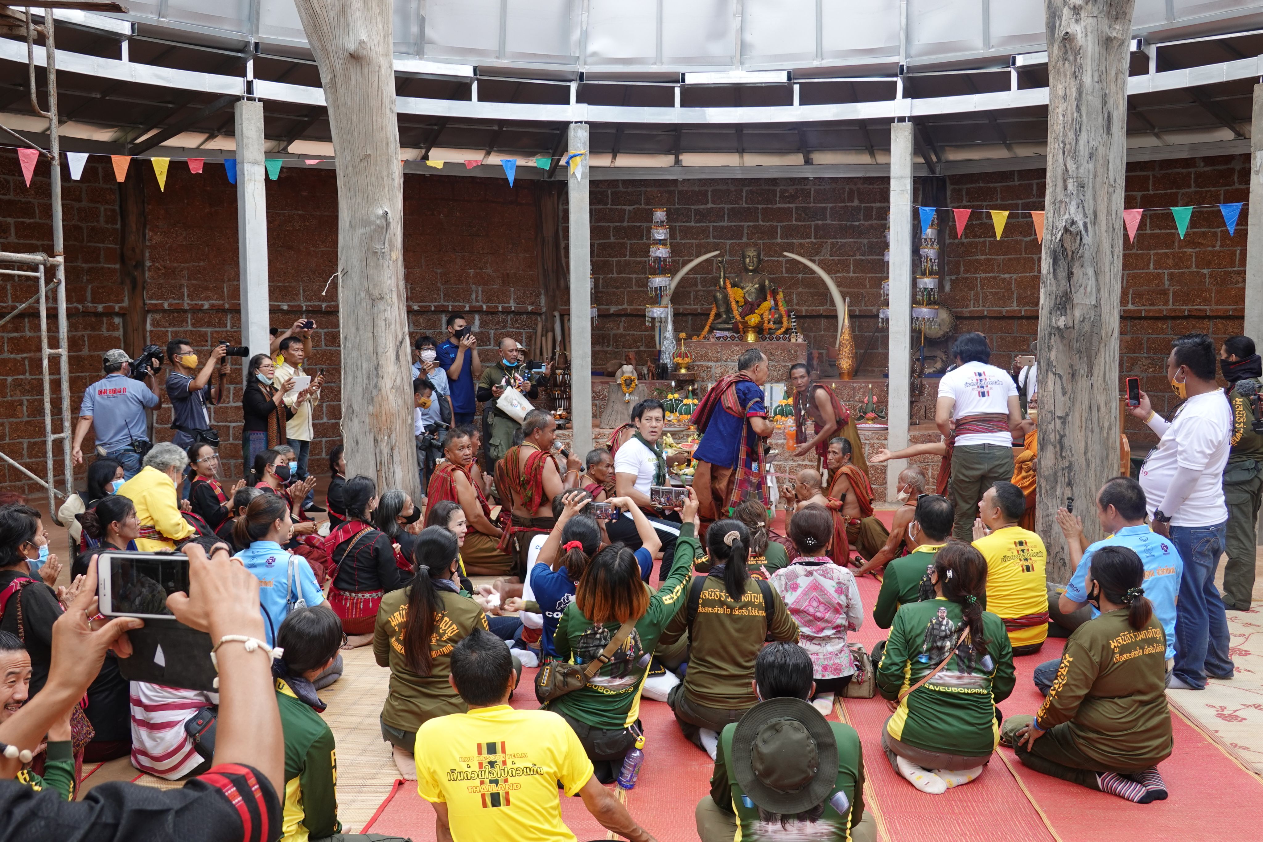 A large group of people sit facing a Pakam shrine. At the front of the group are several hmor ajiang - topless men wearing red cloth draped over their shoulders. At the centre of the shrine is the Pakam god - a gold statue flanked by large ivory tusks on either side. Pakam has yellow floral garlands placed around the statue.  
