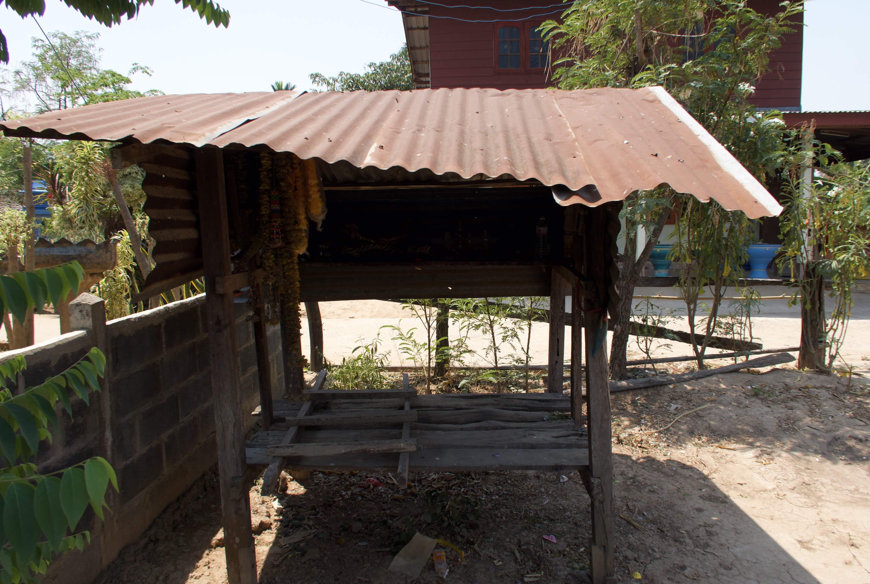 This shrine made of four wooden posts, a wooden base, upper shelf, and a corrugated iron roof looks empty at first. The dark top shelf holds a Pakam rope as well as flower garlands. Old flowers are grey while newer garlands are still bright yellow.