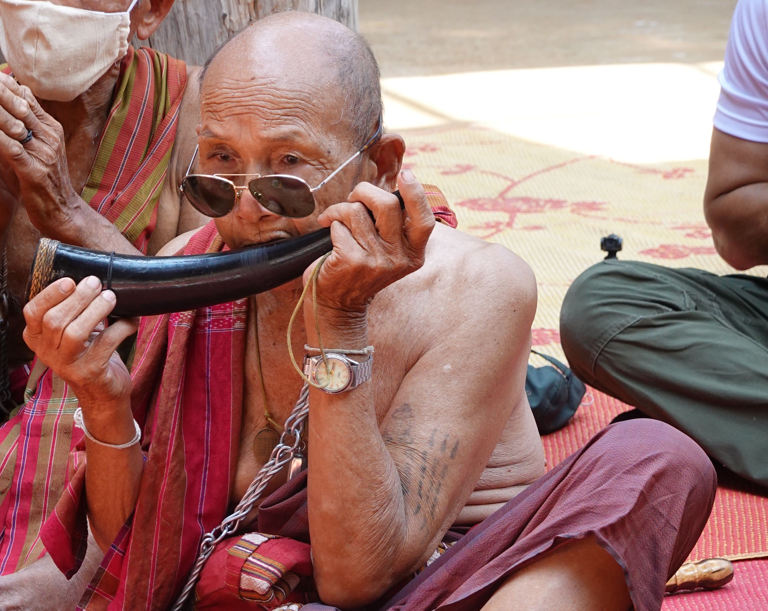 A elderly man sits cross legged, blowing into a curved black horn. He is topless, with a piece of red fabric draped over his shoulder. He has on a pair of aviator sunglasses, a silver watch, and a think iron chain necklace. He has tattoos running down his left arm.