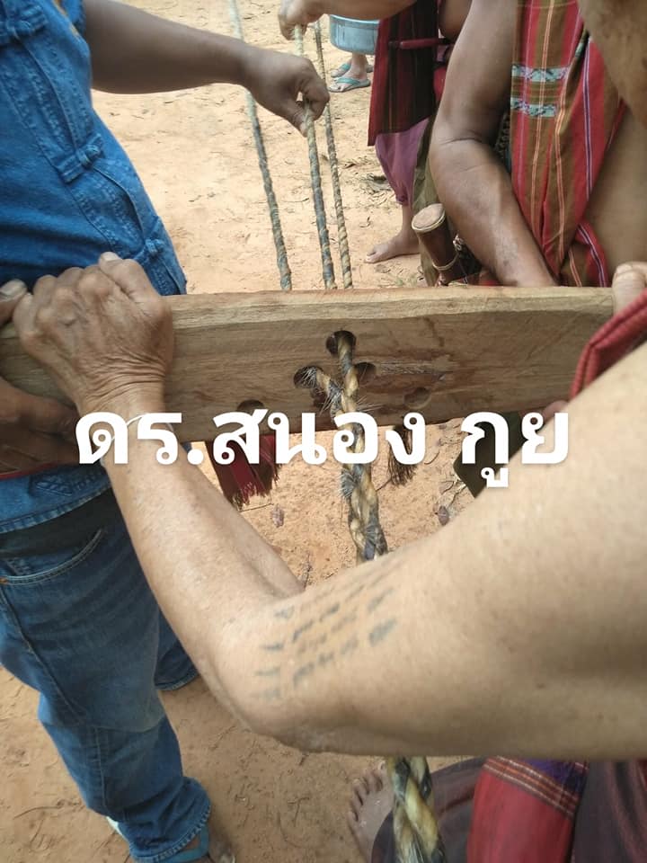 Two men hold up a plank of wood with three holes drilled into it. Three strands of buffalo hide rope are threaded through each of the holes and braided together on the other side of the wooden plank.