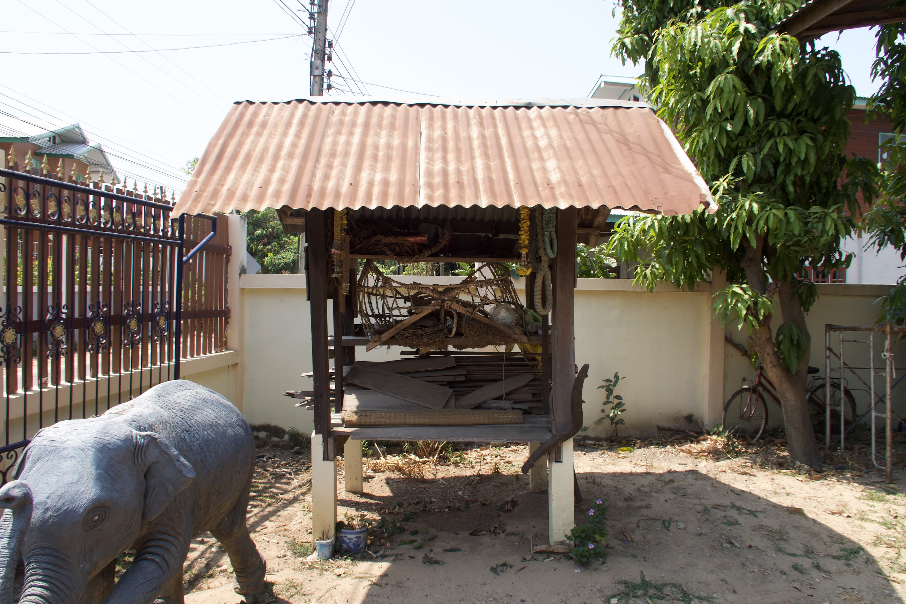 This shrine is a bit older than the other shrines but still has the main four wooden posts and the corrugated iron roof (a bit rusty). At the centre is also a howdah made of wood and rattan bindings. At the base of the shrine are planks of wood and a woven mat. At the top of the shrine is an old, coiled Pakam rope. At the bottom left of the image, in front of the shrine, you can just make out the top of an elephant statue.