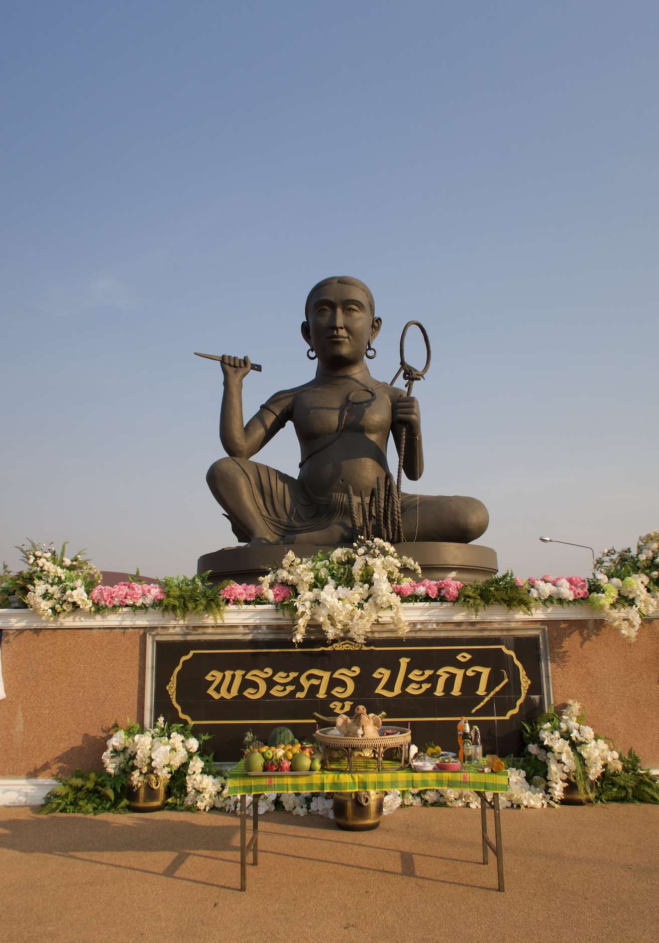 Large, dark grey statue of Pakam. Pakam is a male-presenting figure with a serene expression and dangling earrings. He carries a knife in his right hand and a Pakam rope in his left hand. He is sitting cross legged, with his right knee raised. He is topless, and wearing cloth wrapped around his waist. He is elevated on a platform edged with pink and white flowers. Underneath is a sign that says Phra Khru Pakam in Thai. If on mobile, you will also notice a table under the shrine with various offerings. At the centre is a pig's head.