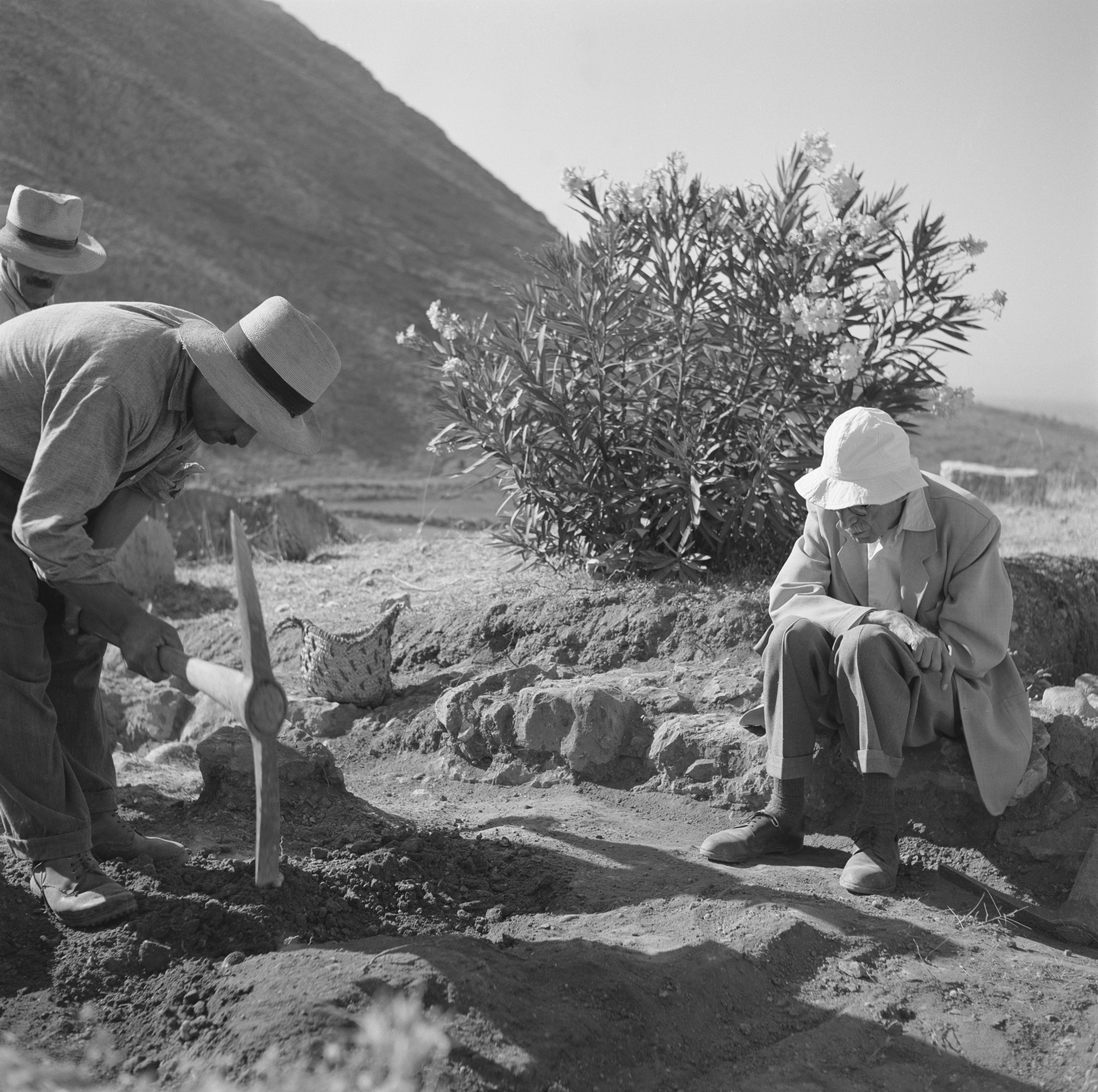 A man digs with a pickaxes as two others look on.
