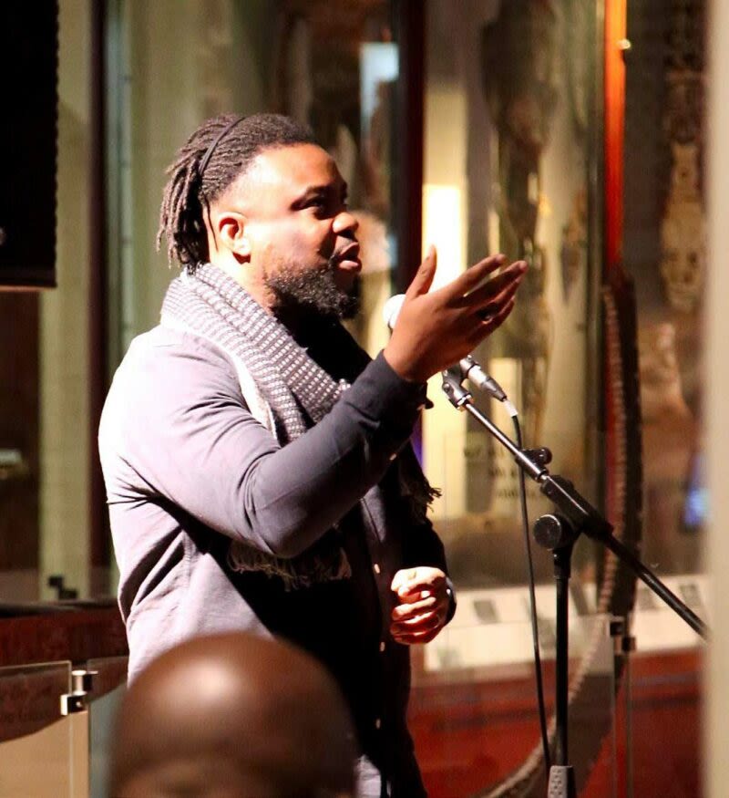 A man speaks into a microphone and raises hand during a performance at the Museum of Archaeology and Anthropology.