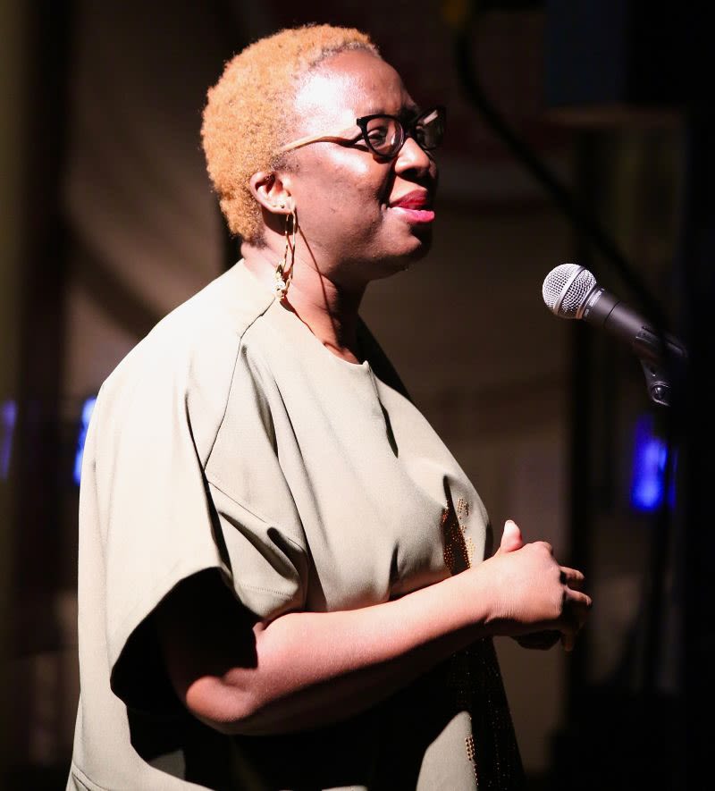 A woman with short blonde hair smiles and speaks into a microphone at the Museum of Archaeology and Anthropology. 