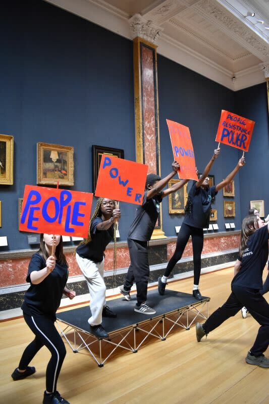 Four young people stand on a small stage and hold out red signs reading 'People' and 'Power'.