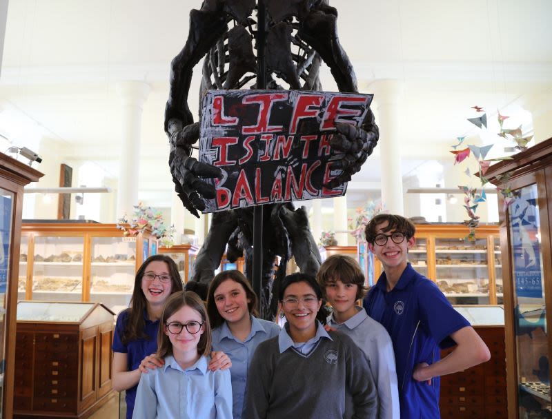 A group of six students smile in front of a skeleton holding a sign reading Life is in the balance at the Sedgwick Museum.