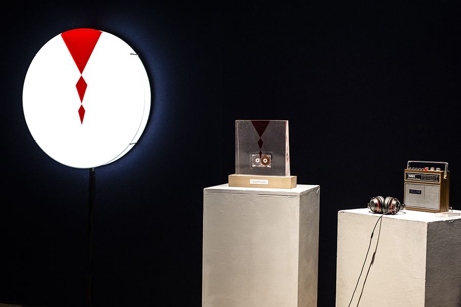 On the left is a bright white light box, with a red motif, in the middle is a transparent acrylic sculpture with the same red motif and a cassette tape embedded within the work, on the right is a cassette player with a pair of headphones.