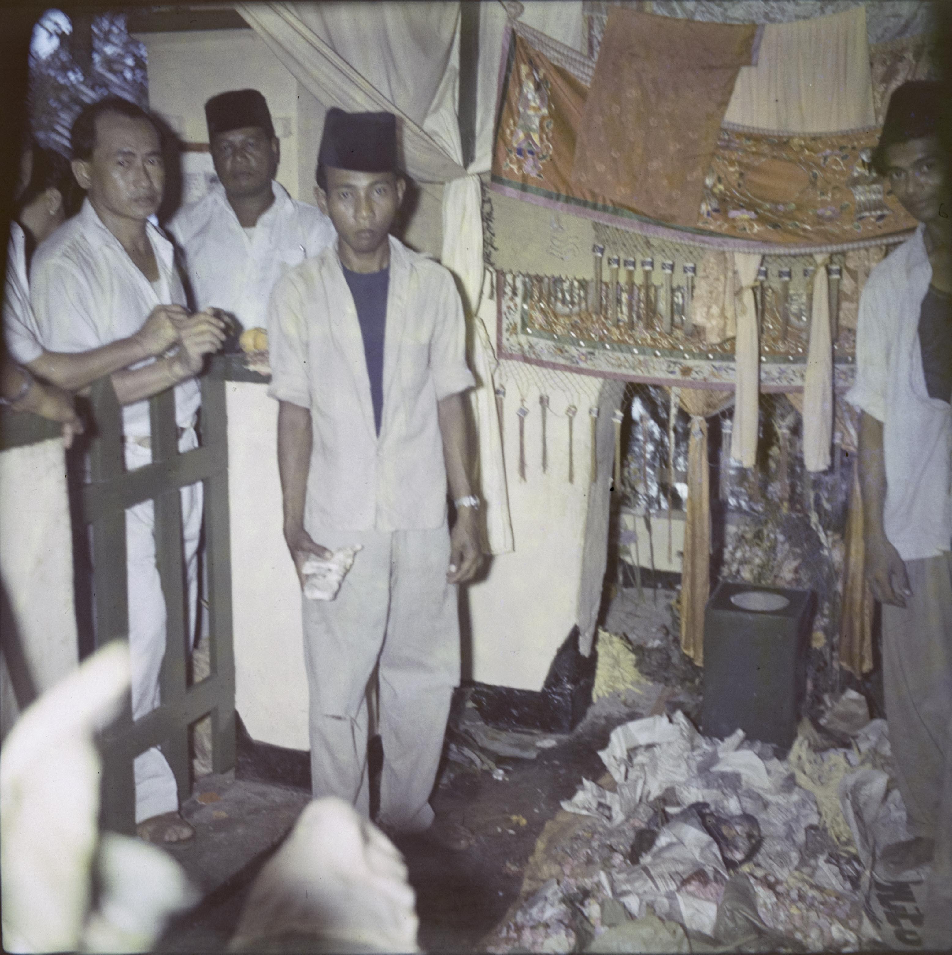 In front of gold and yellow tasselled cloth is a pile of crumpled papers. A young man wearing trousers, short-sleeved shirt, vest and a cloth hat stands holding a similar crumpled paper. Behind him is an older man wearing a black hat and a long white robe, carrying fruit. Two more men lean on a wooden picket fence at the side, looking in.