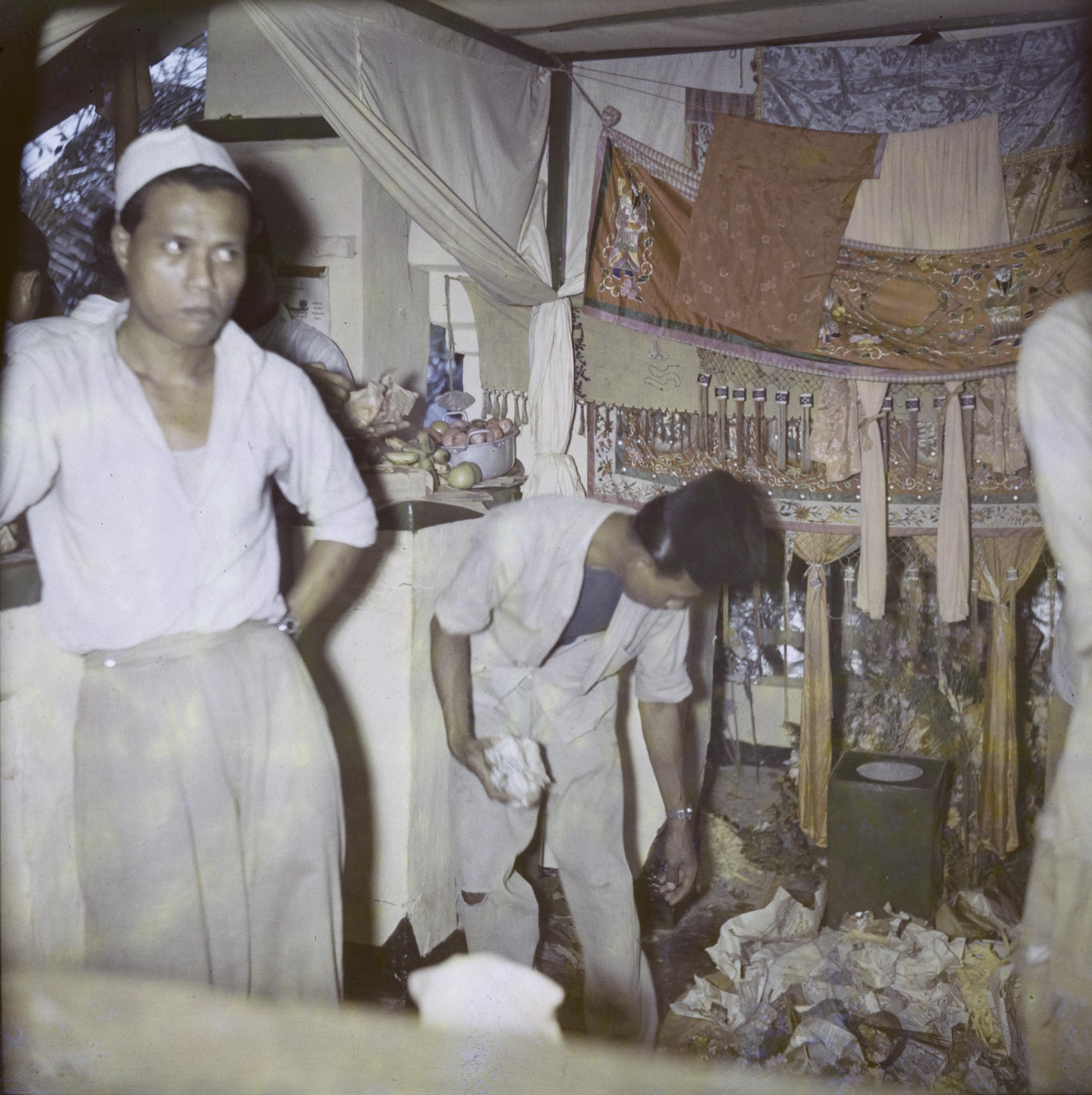 In front of gold and yellow tasselled cloth is a pile of crumpled papers. A young man wearing trousers, short-sleeved shirt, vest and a cloth hat bends down holding a similar crumpled paper. Beside him is a second man in trousers, shirt and white hat, with one hand on his hip and looking upwards. 