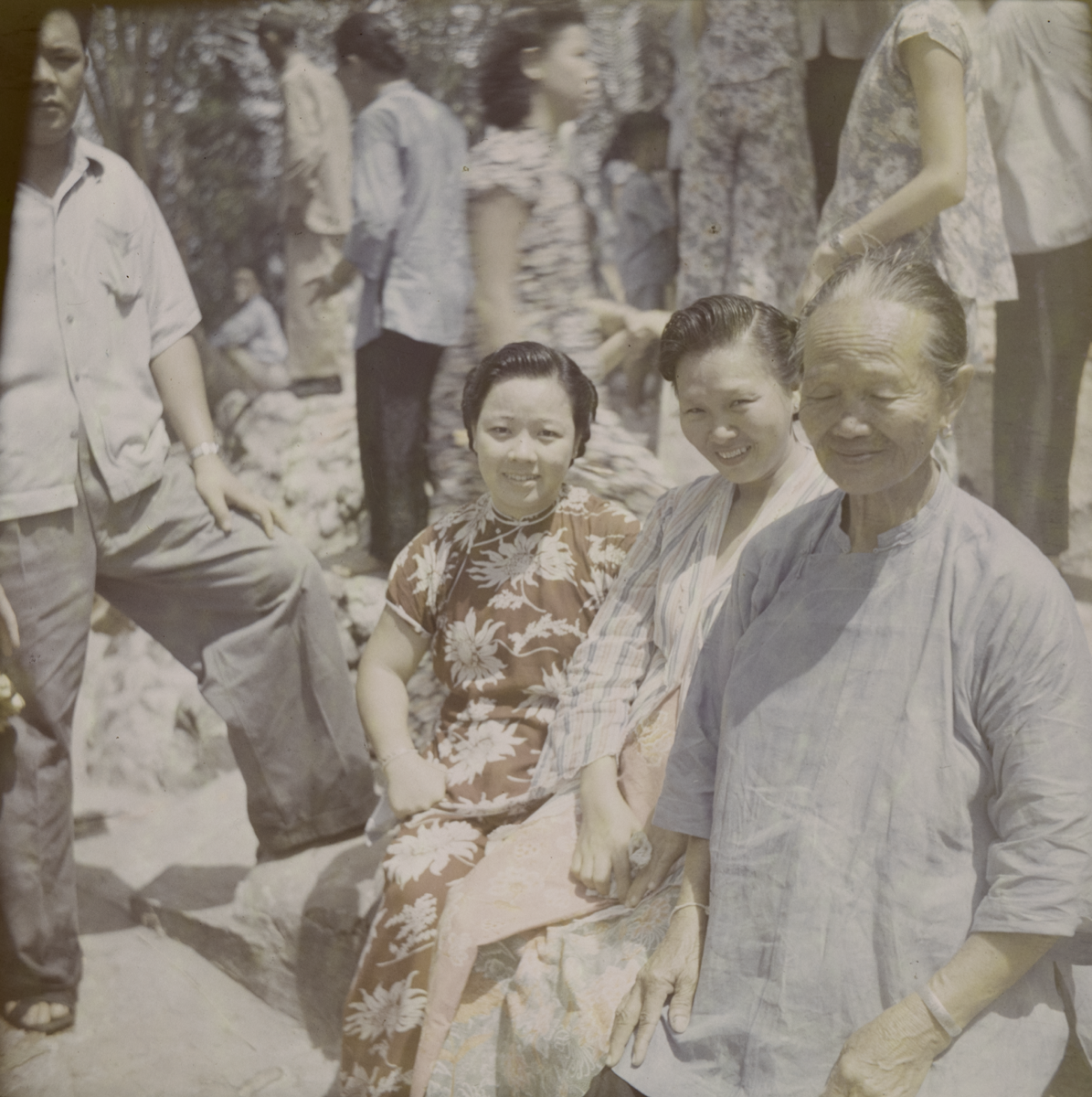 Three Chinese women, one older and two younger sit on a stone wall in an outdoor space with a heap of stones behind where a group of men and women are standing. On the right, the older woman wears a plain blue collarless blouse, buttoned at the side and has her hair tied back. Beside her, a possibly pregnant woman wears a striped shirt and a pastel coloured print skirt. On the left is a younger woman wearing a cheongsam printed with flowers. Next to the women is a man in a short-sleeved shirt and trousers, standing with his left leg on the wall.