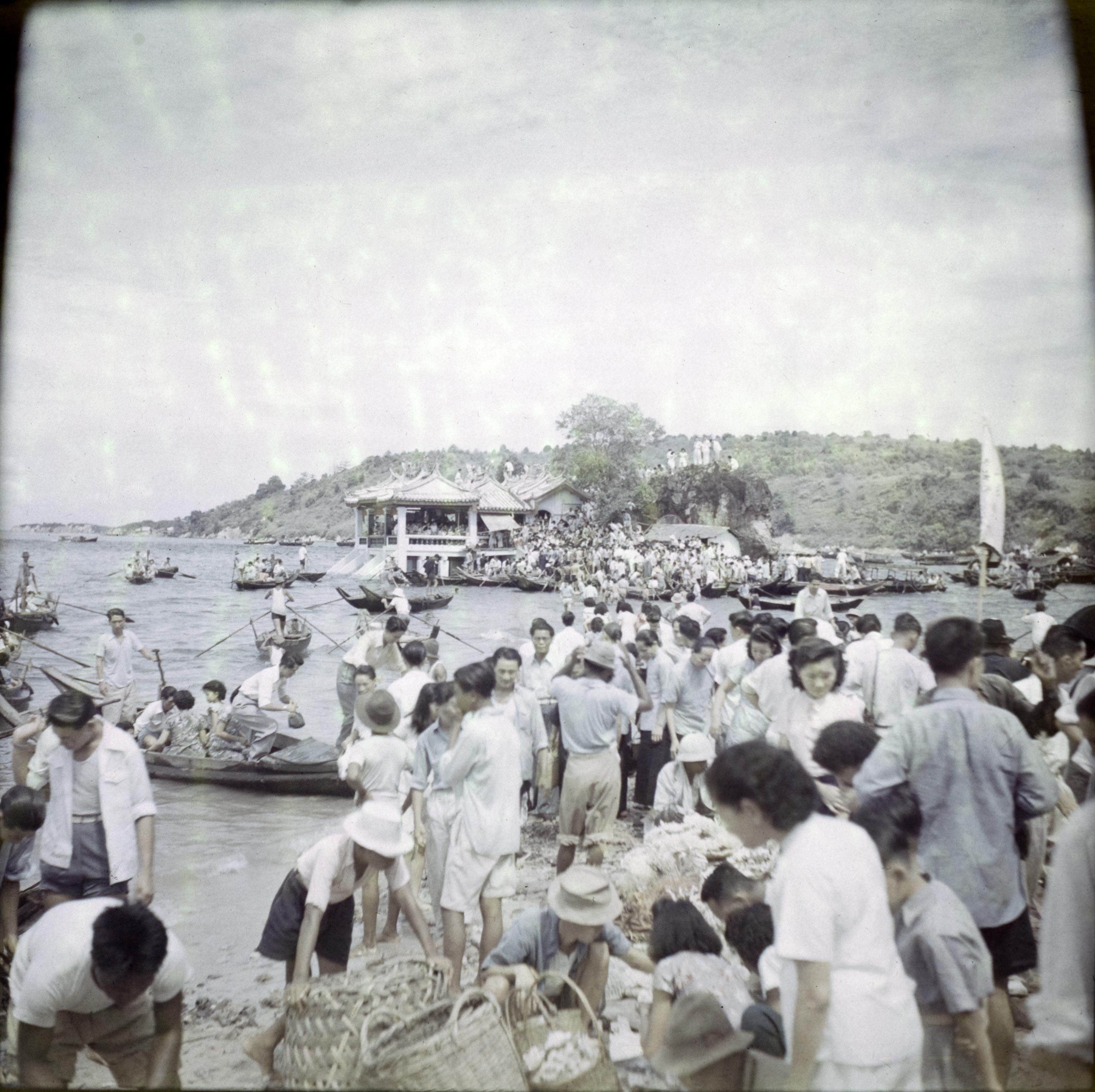 On the seashore, women and children lean over baskets of corals. There are several small rowing boats in the water close to shore, and a building visible around the bay.