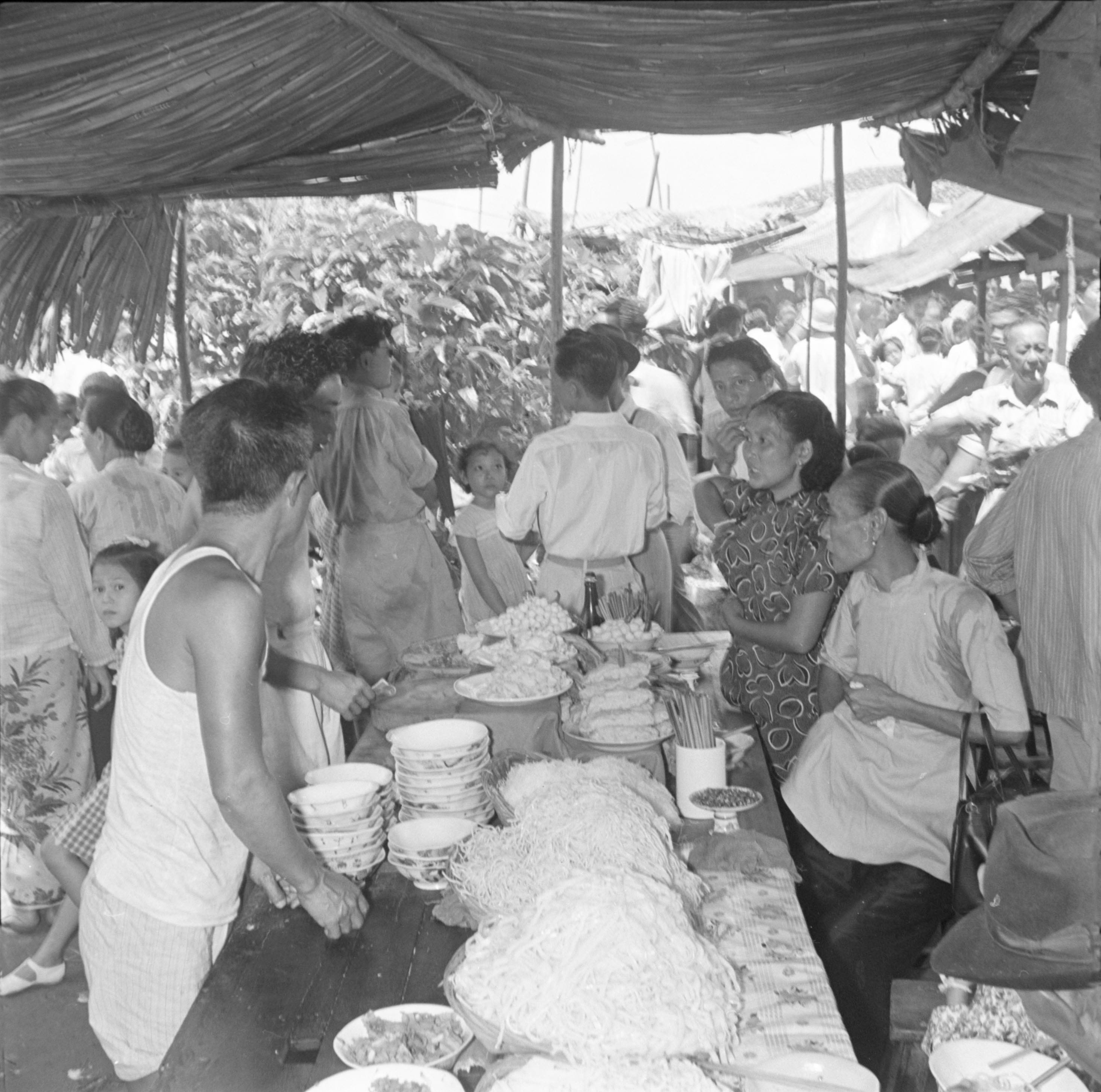 Under a canopy of leaves, a long table is laid with large dishes of noodles and other food, and stacks of rice-bowls. On the right two women sit at the table, while a group of men stand to the left.
