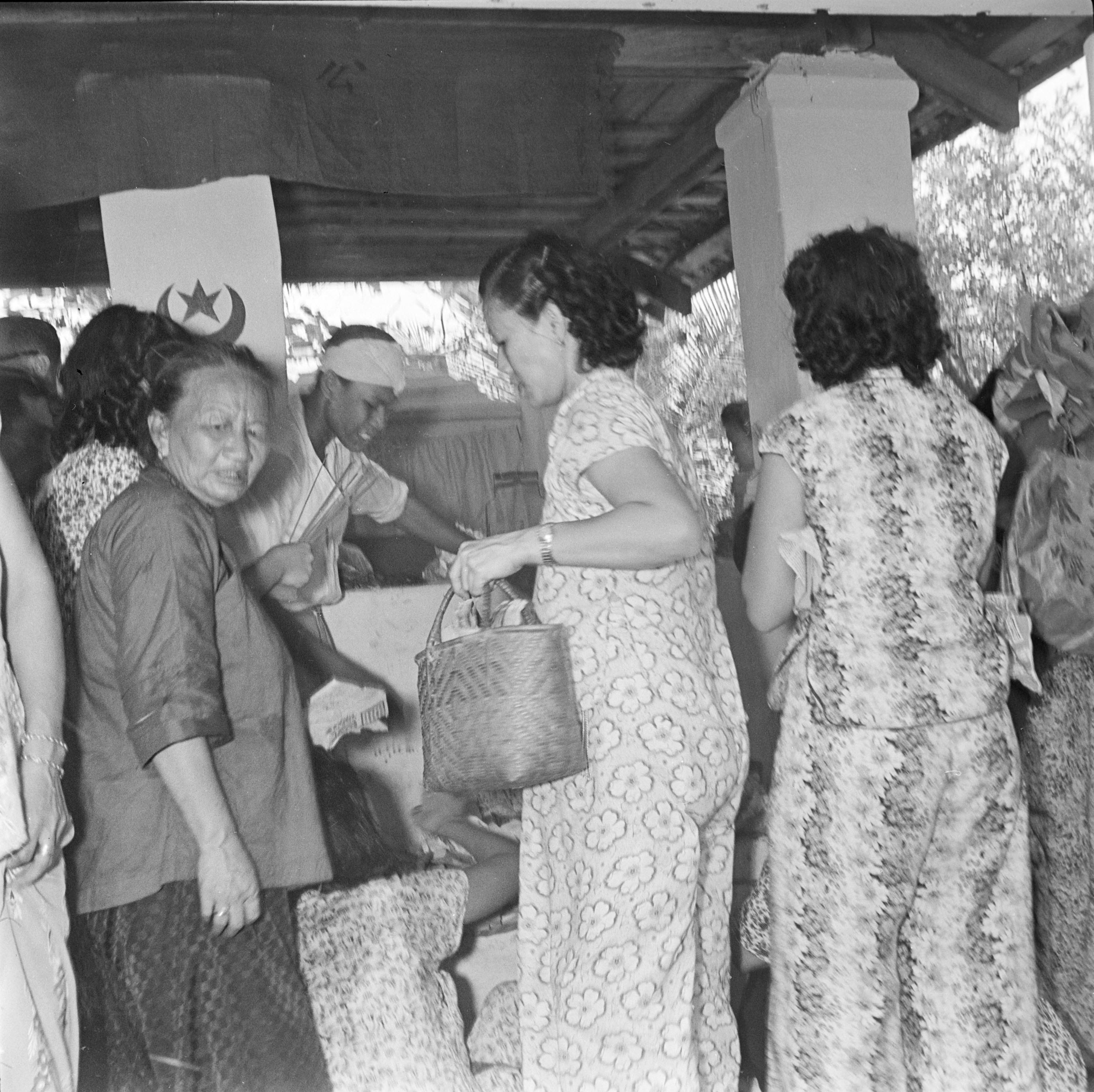 A small group of Chinese women stands under the roof of a small building with whitewashed walls. On one wall is painted a crescent moon and star symbol. 