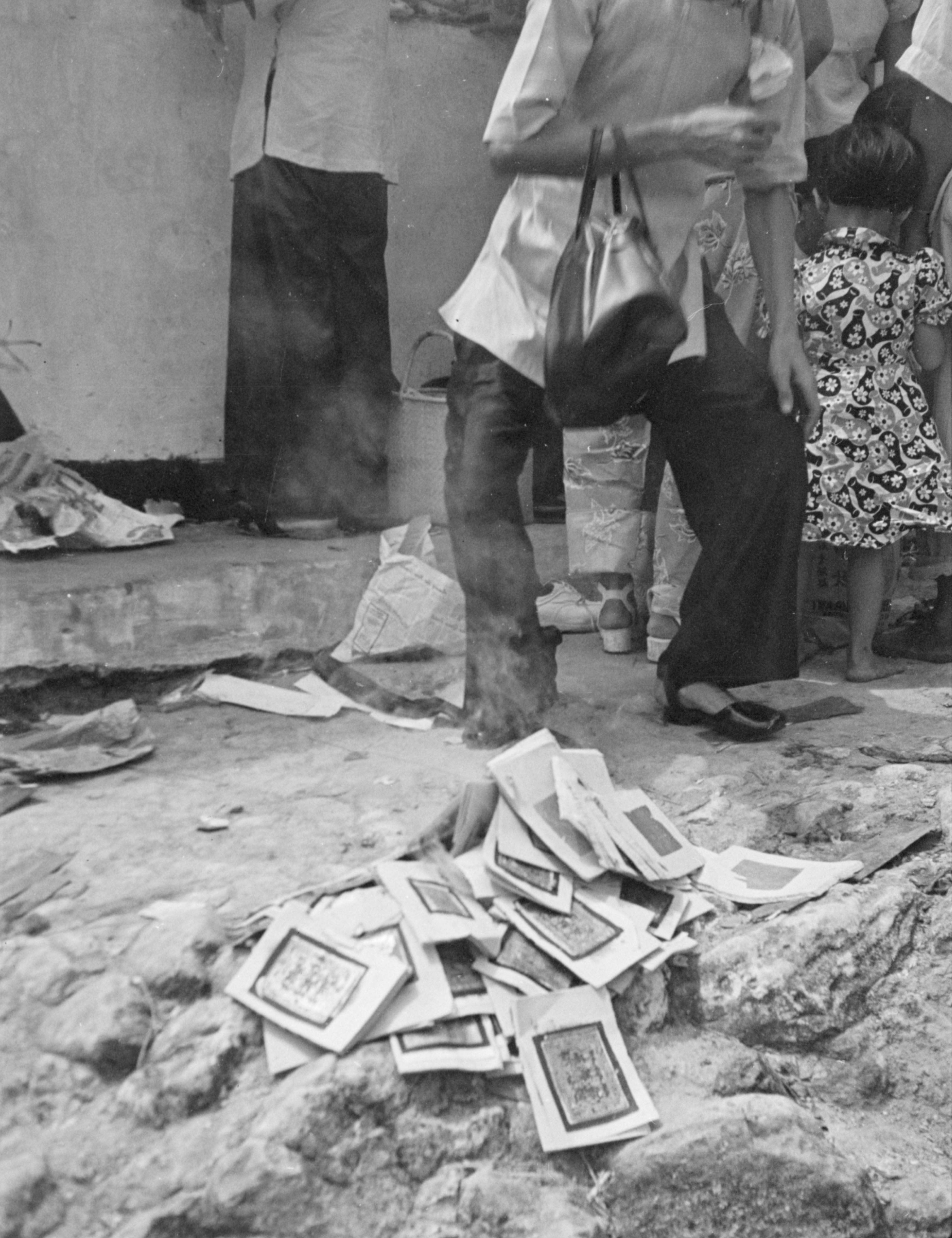 In the centre front, on the ground is a heap of printed fu (符, charm papers) with wisps of smoke coming from them. Behind the papers, an older woman (cropped by the frame) stands wearing dark trousers and a blouse with a handbag over her left arm. 