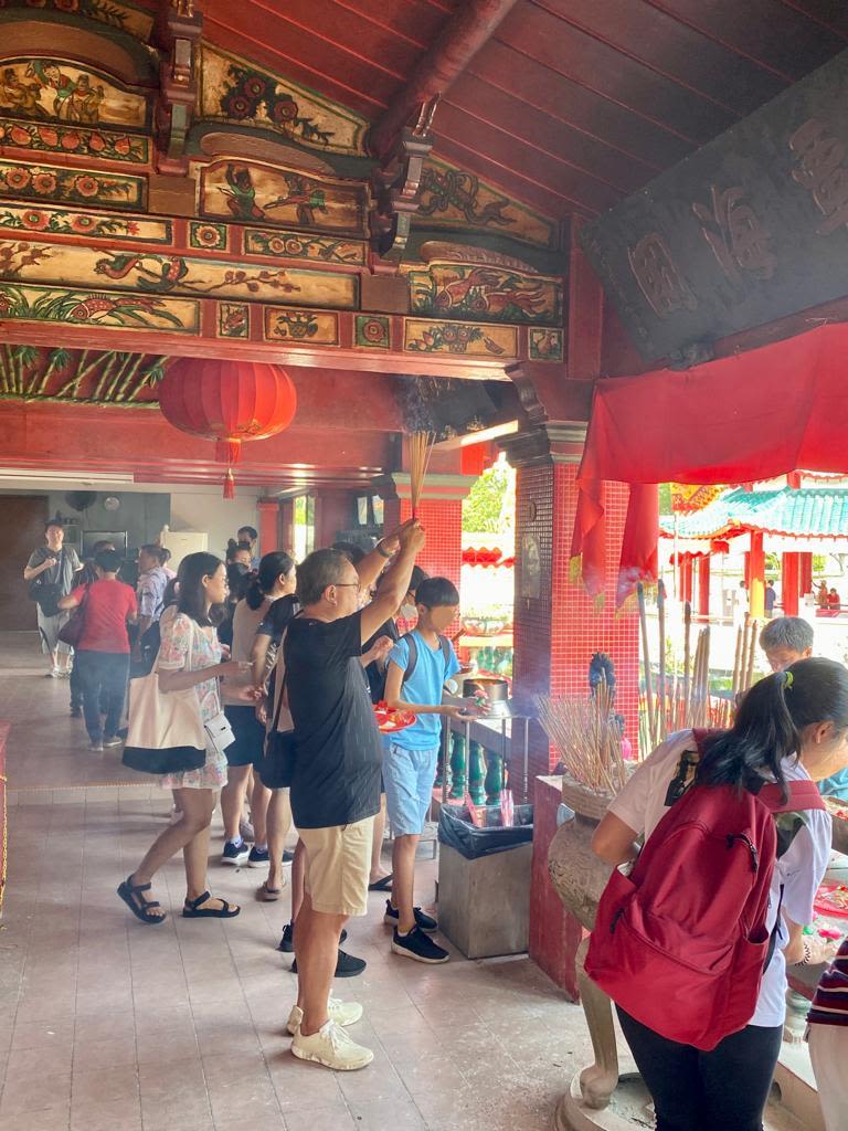 Men, women and children holding joss sticks, facing out of a Chinese temple with carved and painted walls and ceilings.  