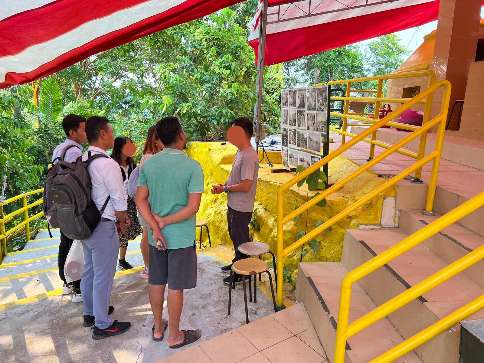 A man stands in front of a display of photographs, speaking to a group of five men and women.