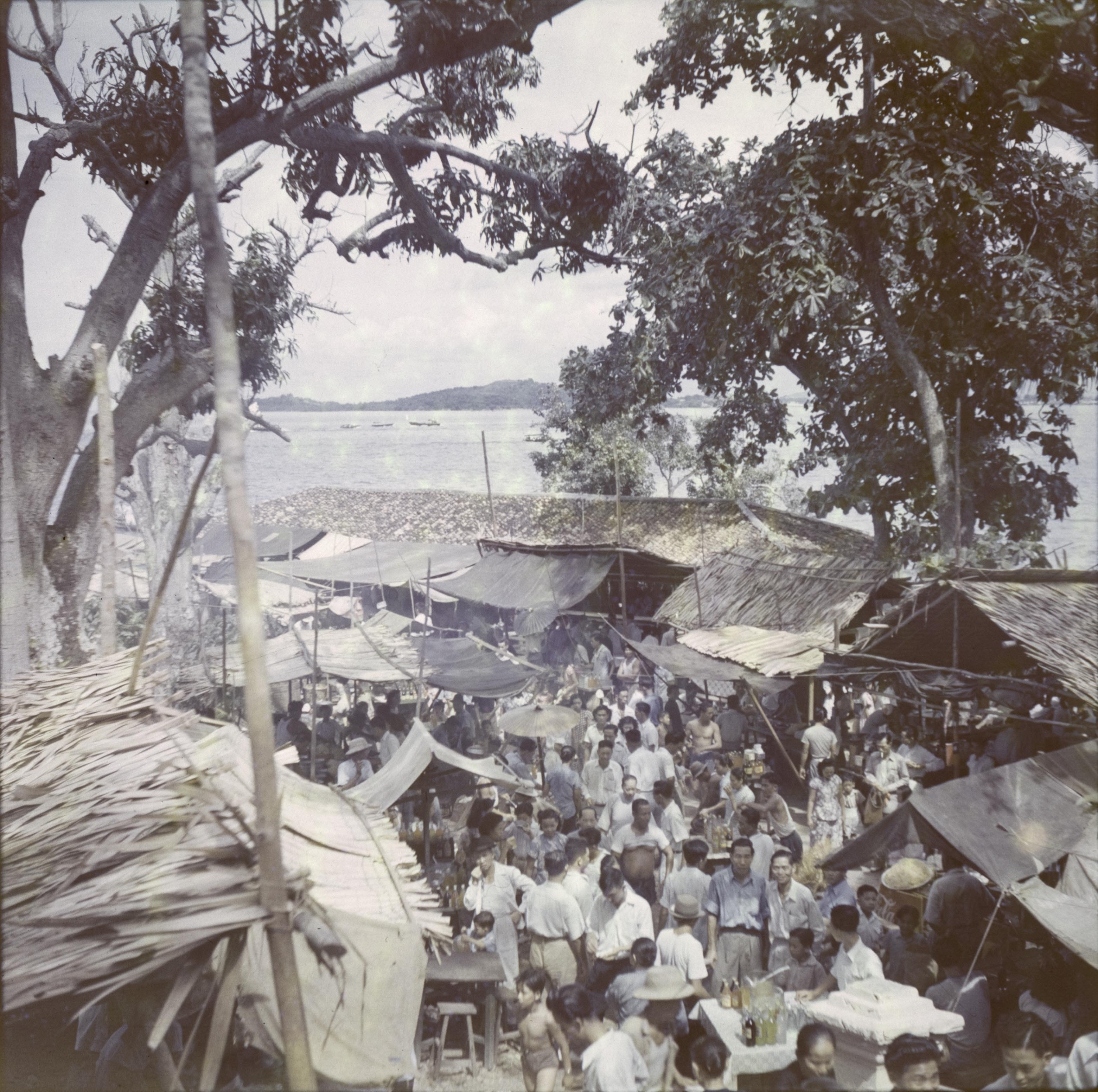 A large crowd walk along a street lined with stalls and buildings with attap roofs. The sea and another island can be seen above the buildings. 