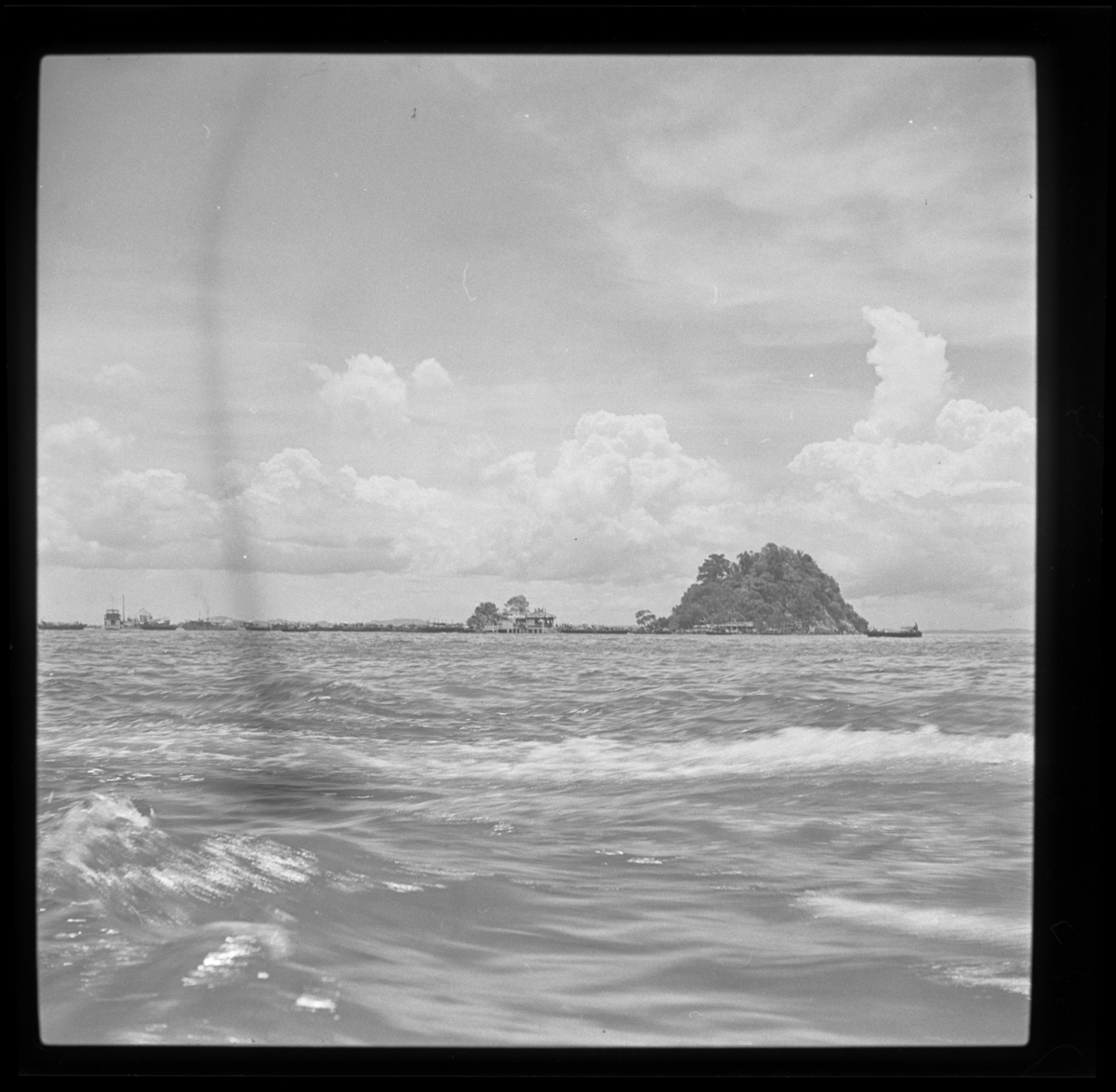 Seen from the sea, on the horizon is a small, low-lying island with a higher clump of trees in the centre right.