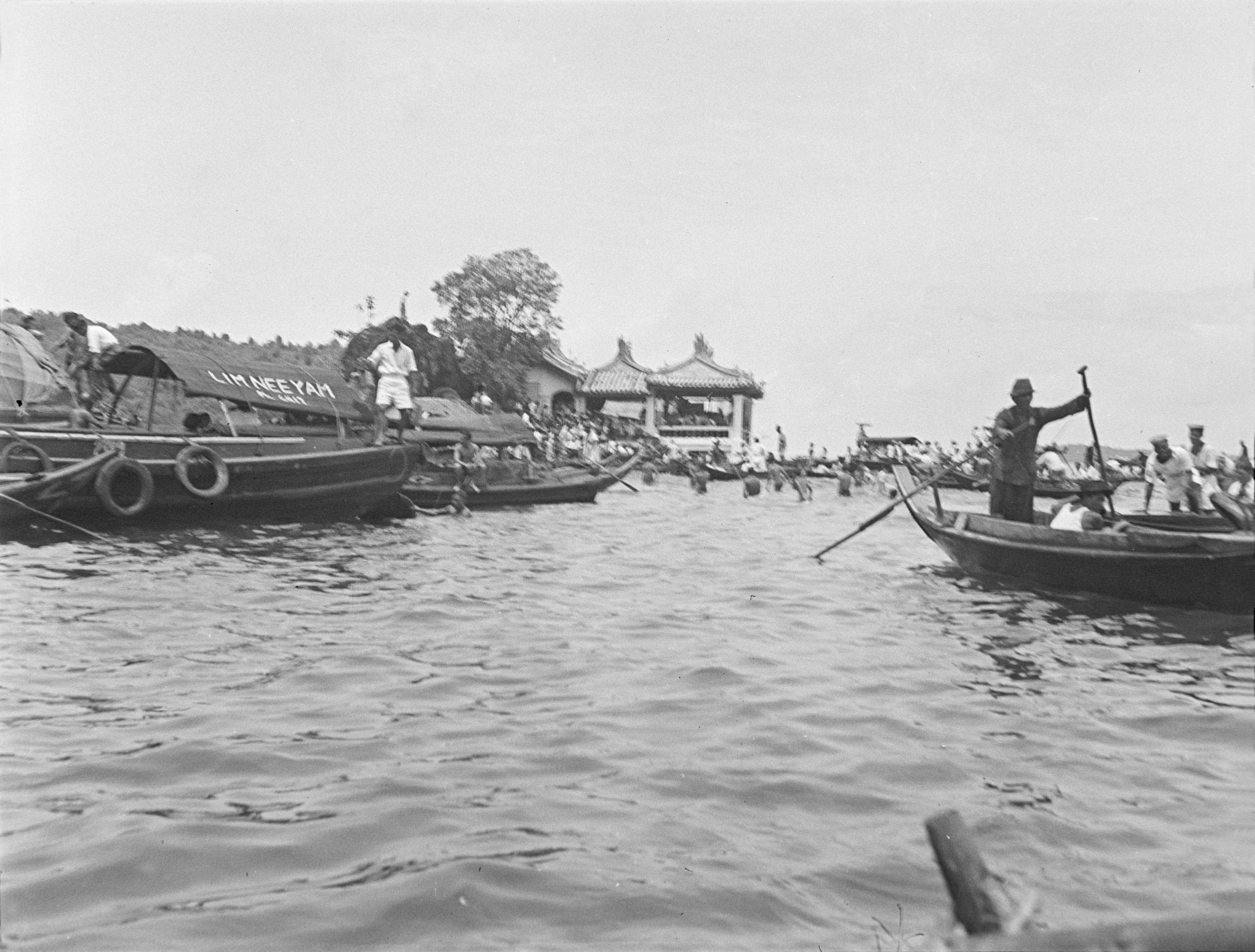 Viewed from the water is a single-storey white building, with tiled roofs. A row of boats is moored by the shore while others are being rowed away to the right. 