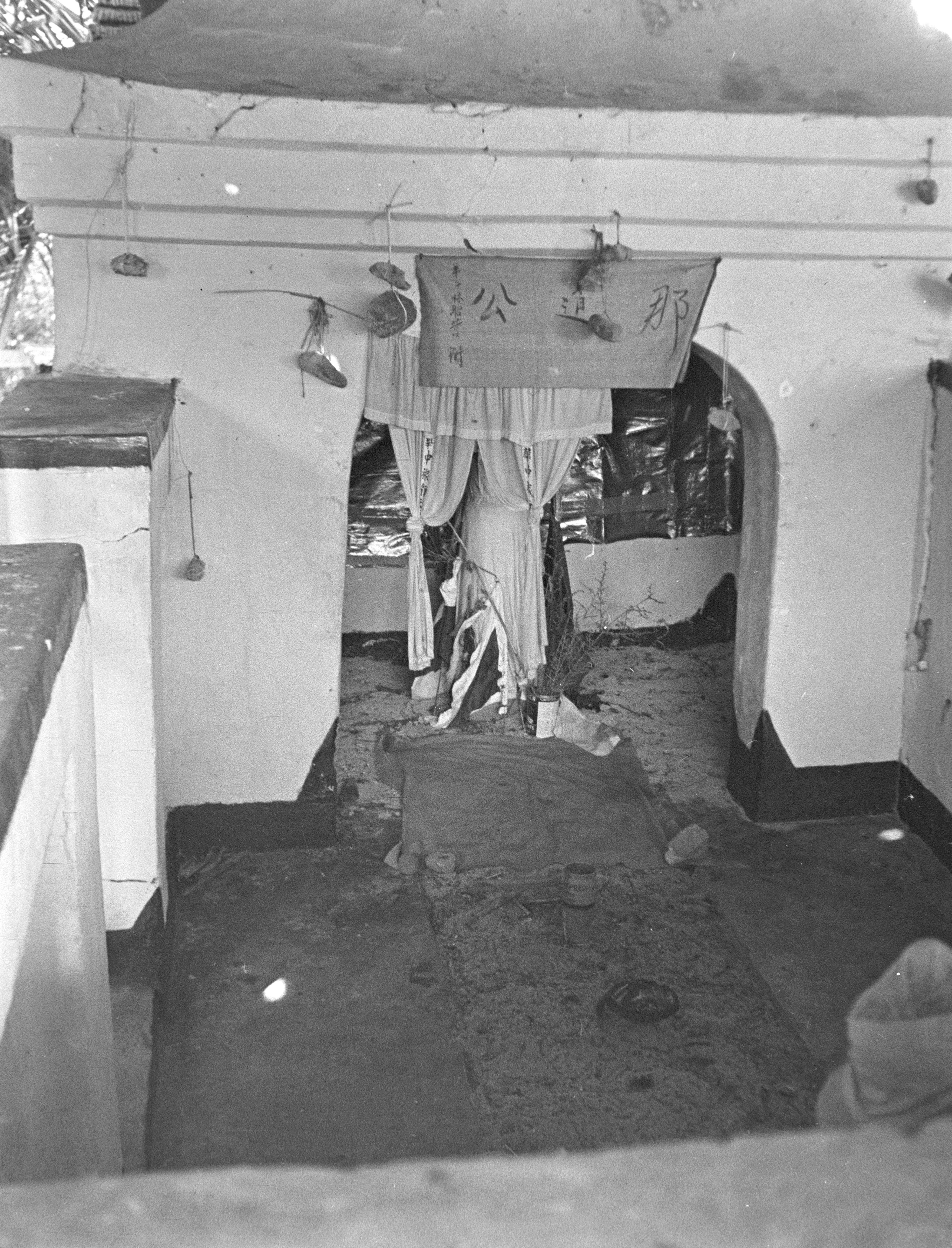 Front view of a shrine. A cloth banner reads 那道公 in Chinese characters, which translates to Datok Kong. Stones are tied with string to the top of the shrine. An oblong stone wrapped with a piece of cloth sits in the middle of the shrine. A curtain with strips of fabric decorated with Chinese characters hang above the stone figure.