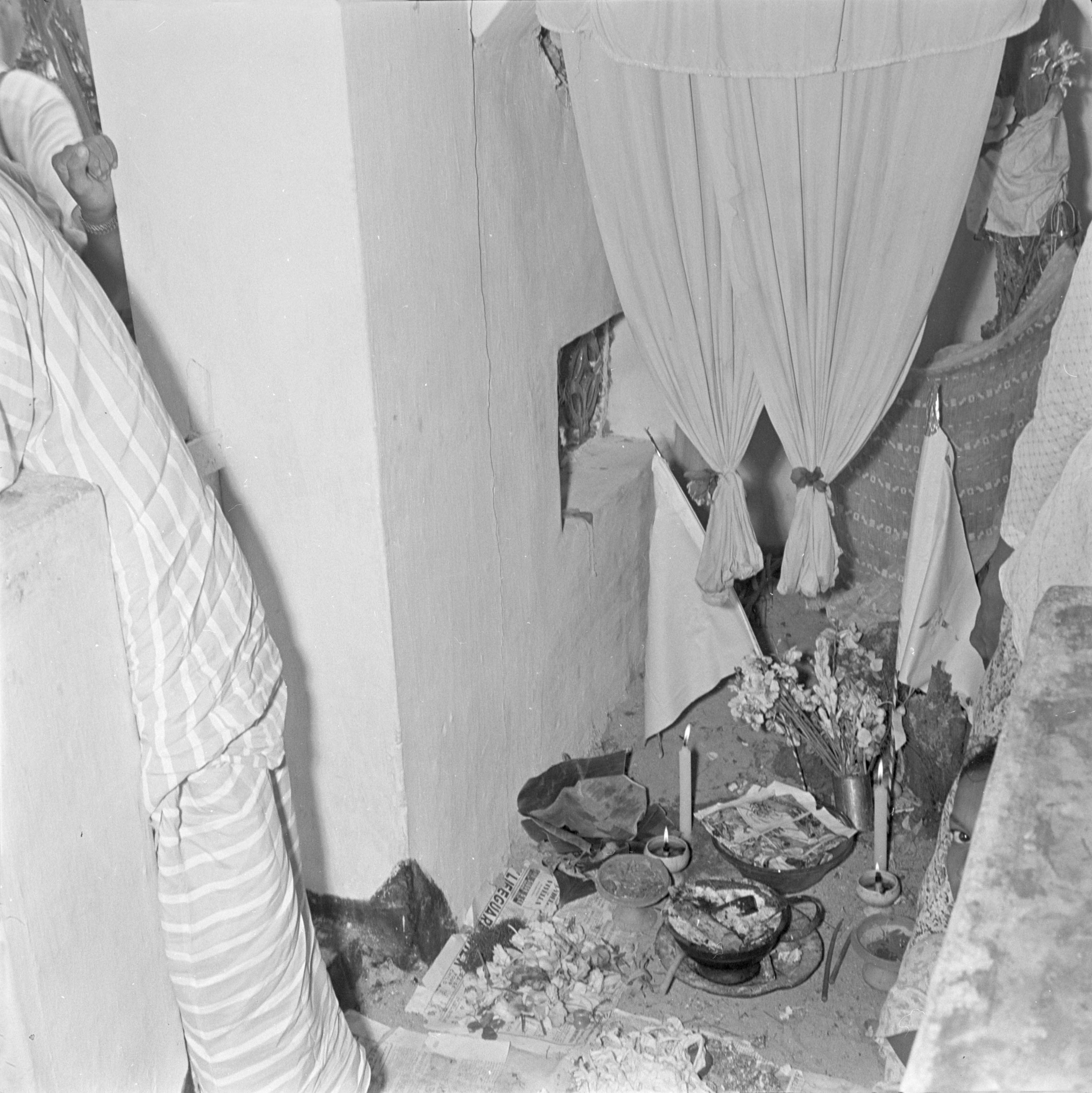 Small offering dishes, candles, flowers and flags have been placed on an earth floor by a whitewashed wall. Two small white curtains hang above the offerings.