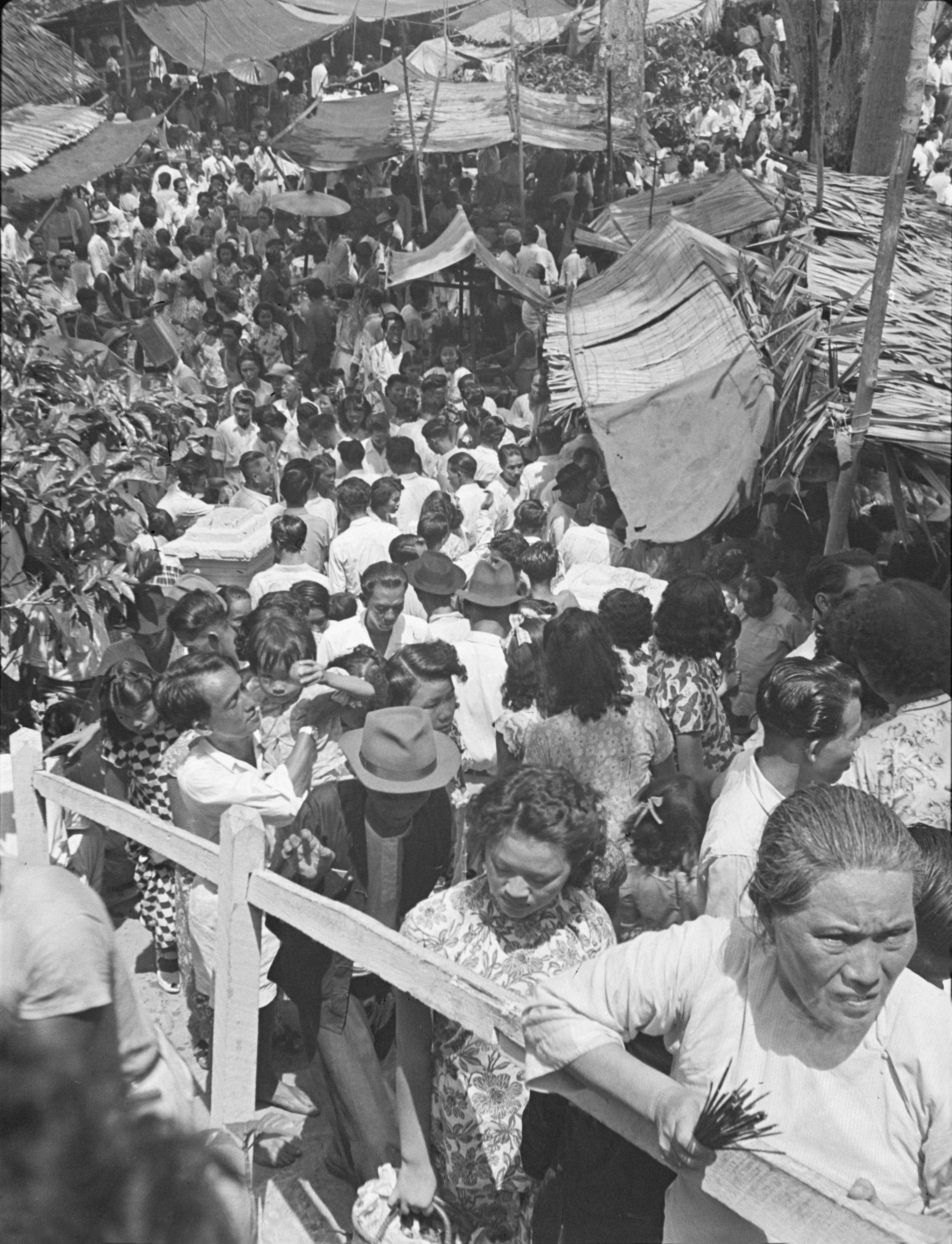 A large crowd of men, women and children walk along a street on either side of which are wooden stalls with canvas roofs. In the front right, a woman leans on a wooden rail, holding a bundle of incense sticks in her right hand. Behind her, a woman carries a basket.