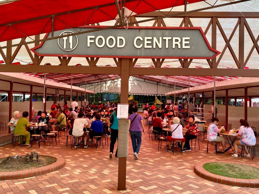 A signboard that reads 'Food Centre'. Round tables with stools in the background, where people gather with food and drinks.