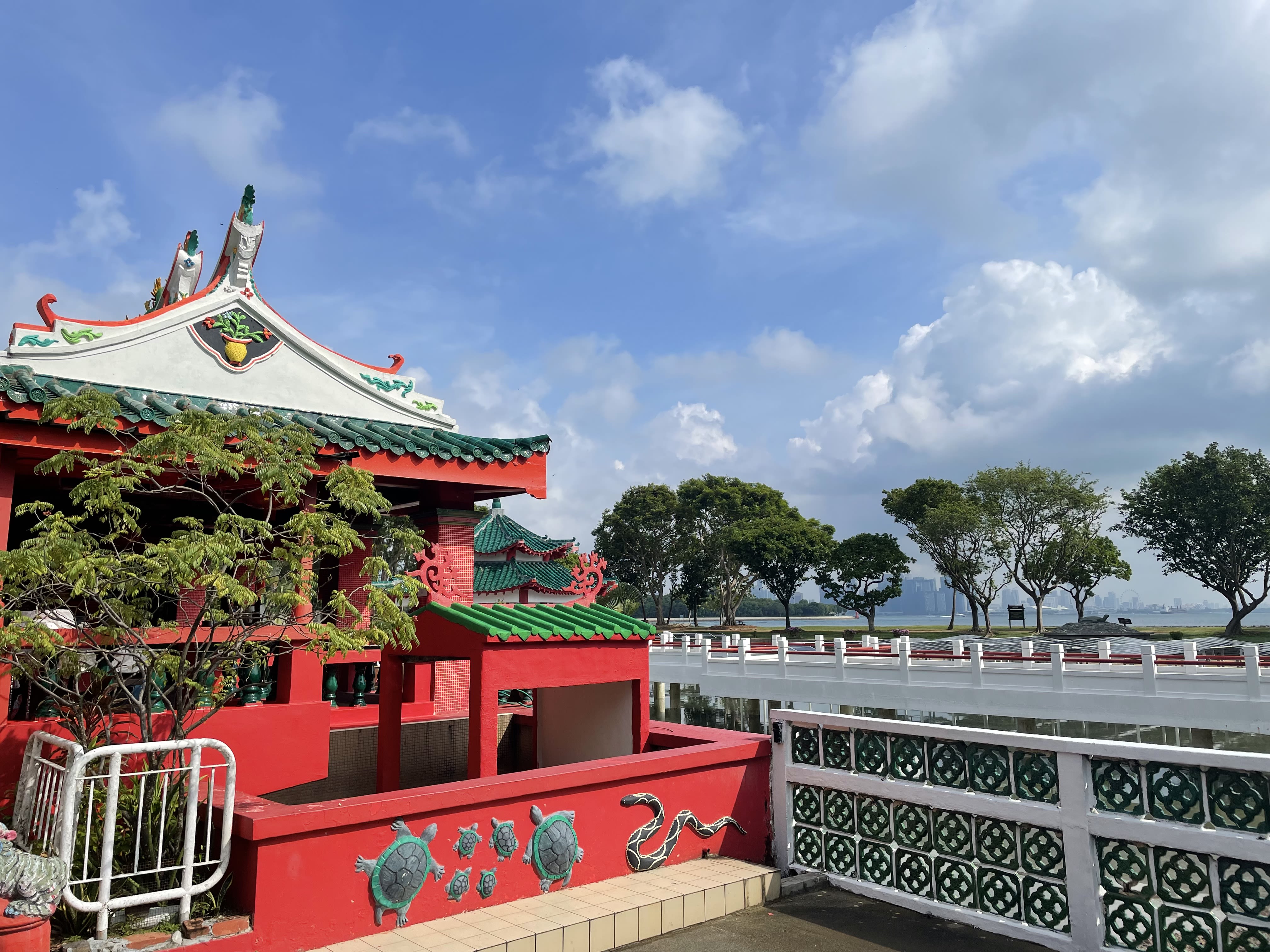 A red, green and white Chinese-style temple with ornate carvings. In the background is the sea and the skyline of a city.