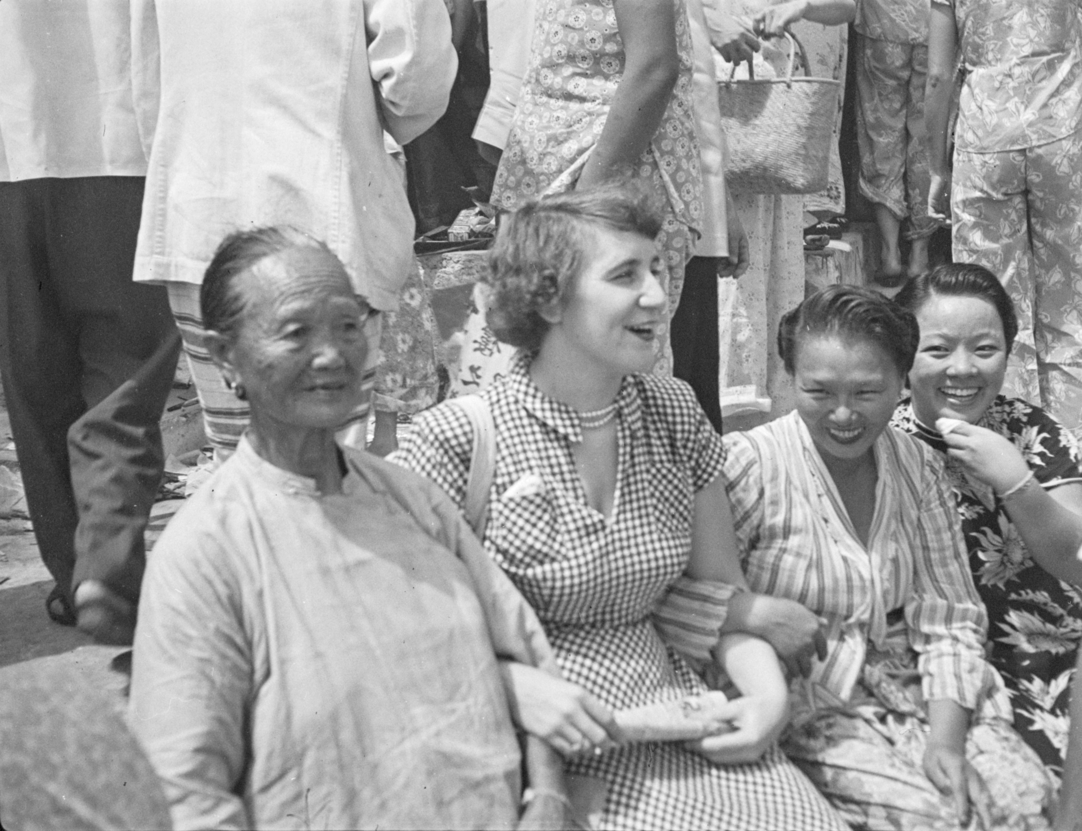 Four women sit, smiling, together. In the centre a European woman, possibly Muriel Ann Elliott, in a check patterned dress and pearls, sits arm in arm with an older Chinese woman on her left and a younger Chinese woman on her right.