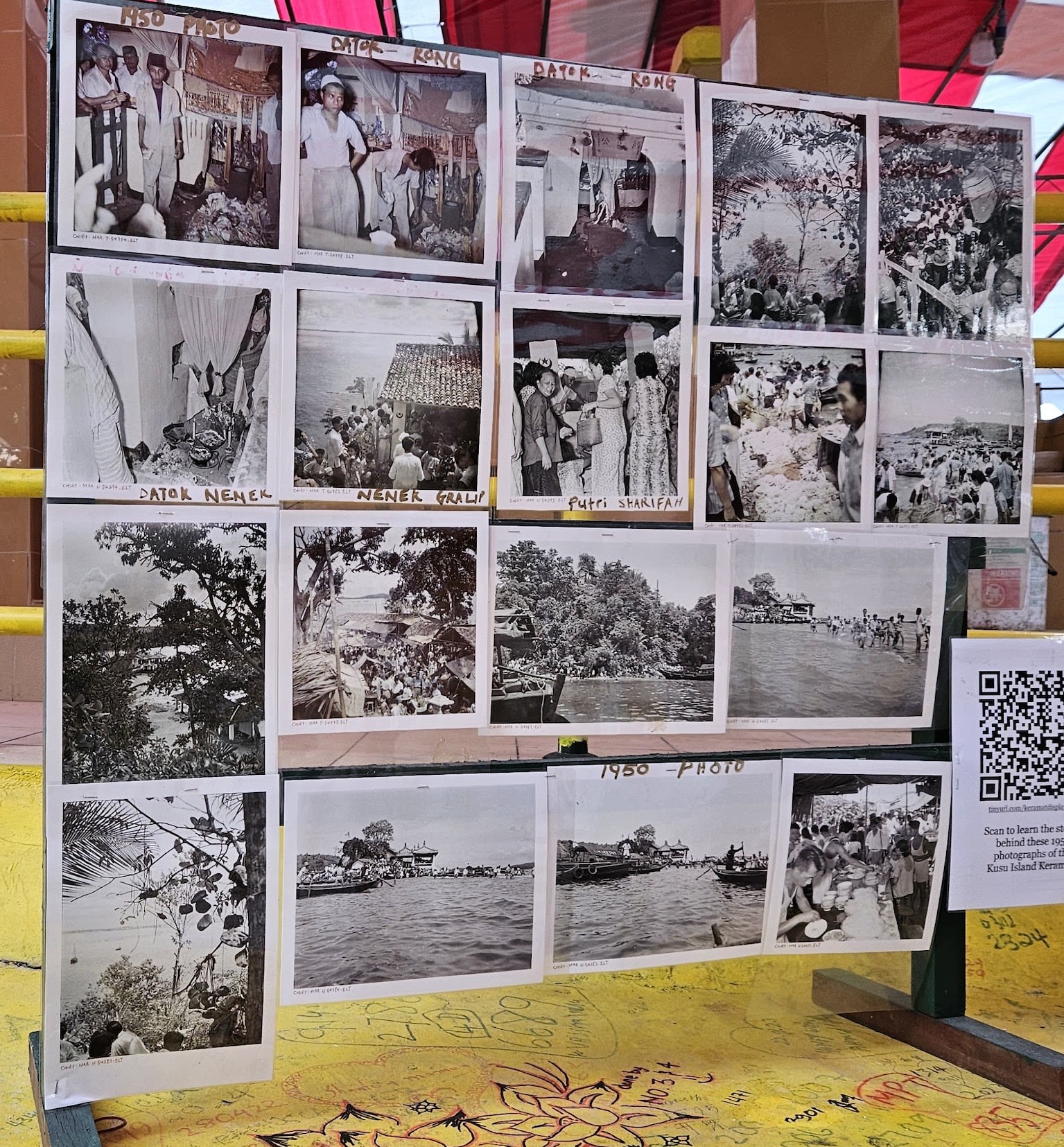 Eighteen printed photographs arranged on a metal stand, placed on a yellow low wall. Some of the photos are labelled with '1950 Photo', 'Datok Kong', 'Nenek Gralip', 'Datok Nenek', and 'Putri Sharifah'.
