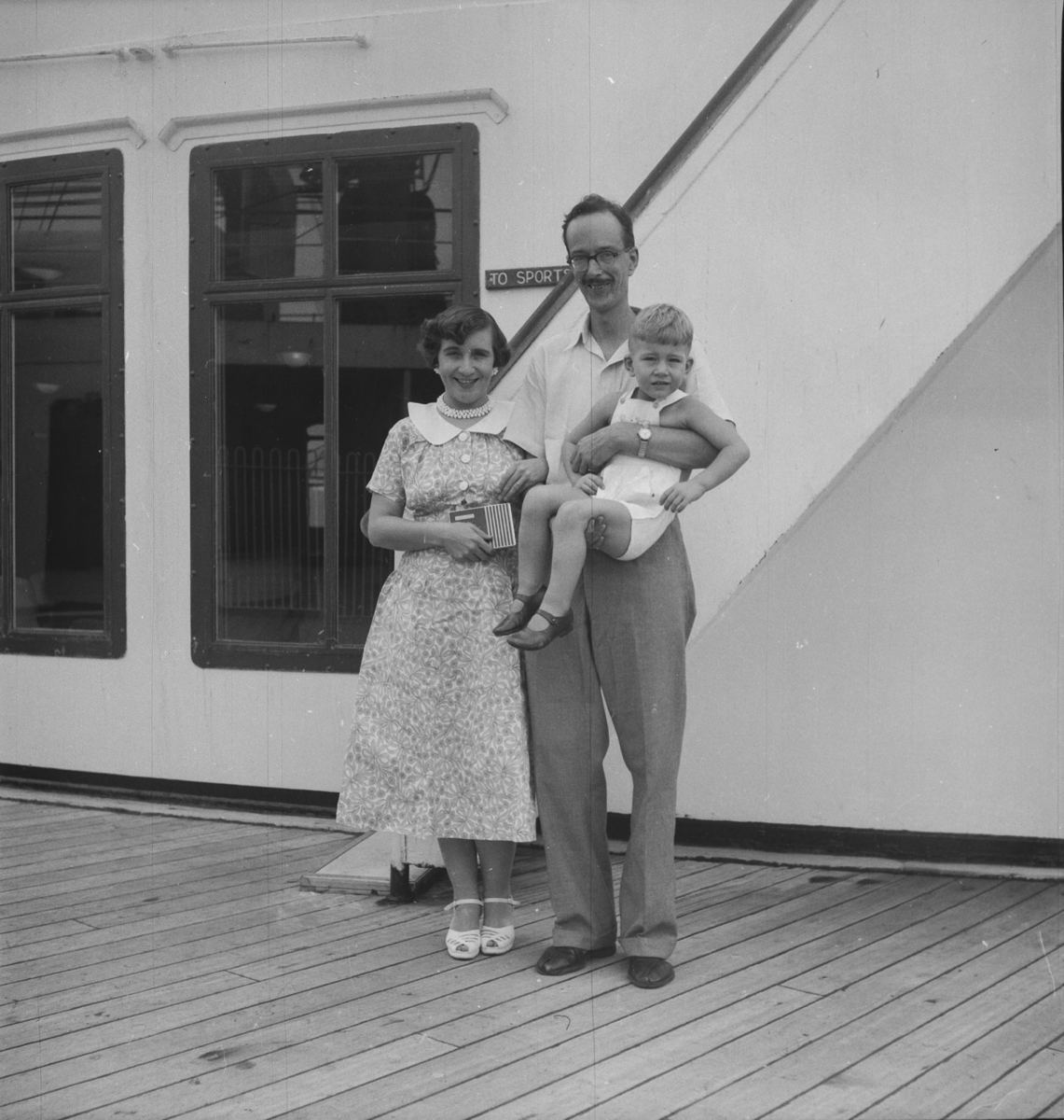 A European man carrying an infant child, with a European woman on his right, standing on the deck of a ship.