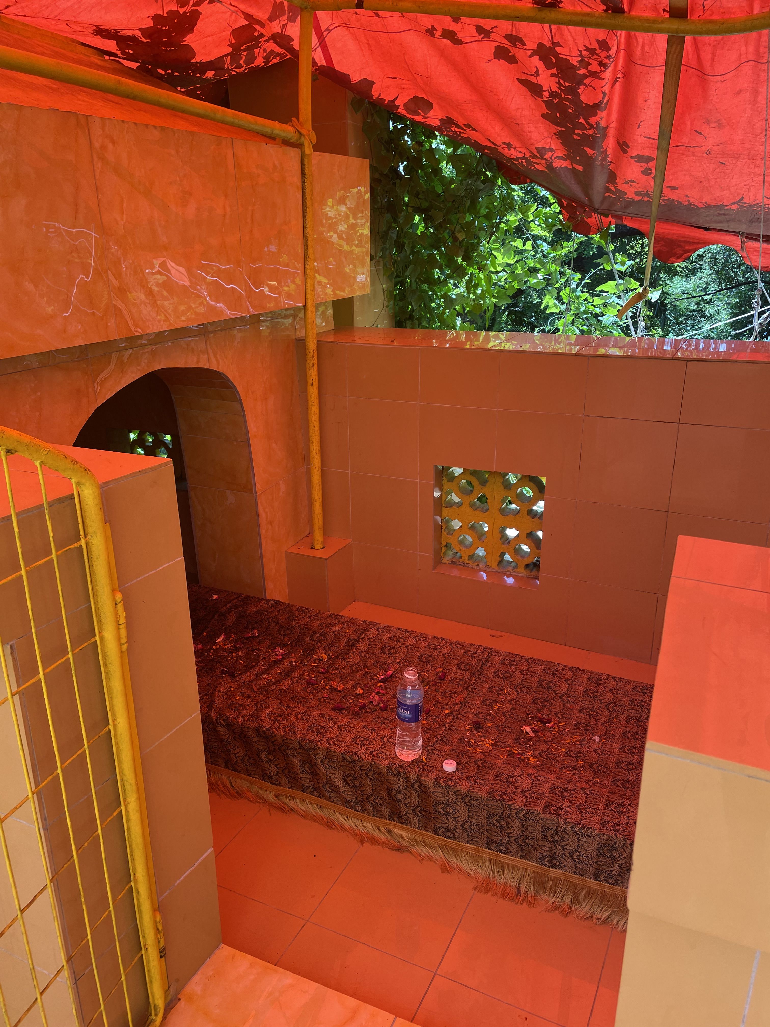 An opened bottle of water placed on top of a cloth covering a rectangular structure on the floor of a yellow shrine. Behind the bottle perforated cement tiles peek through newer tiles.