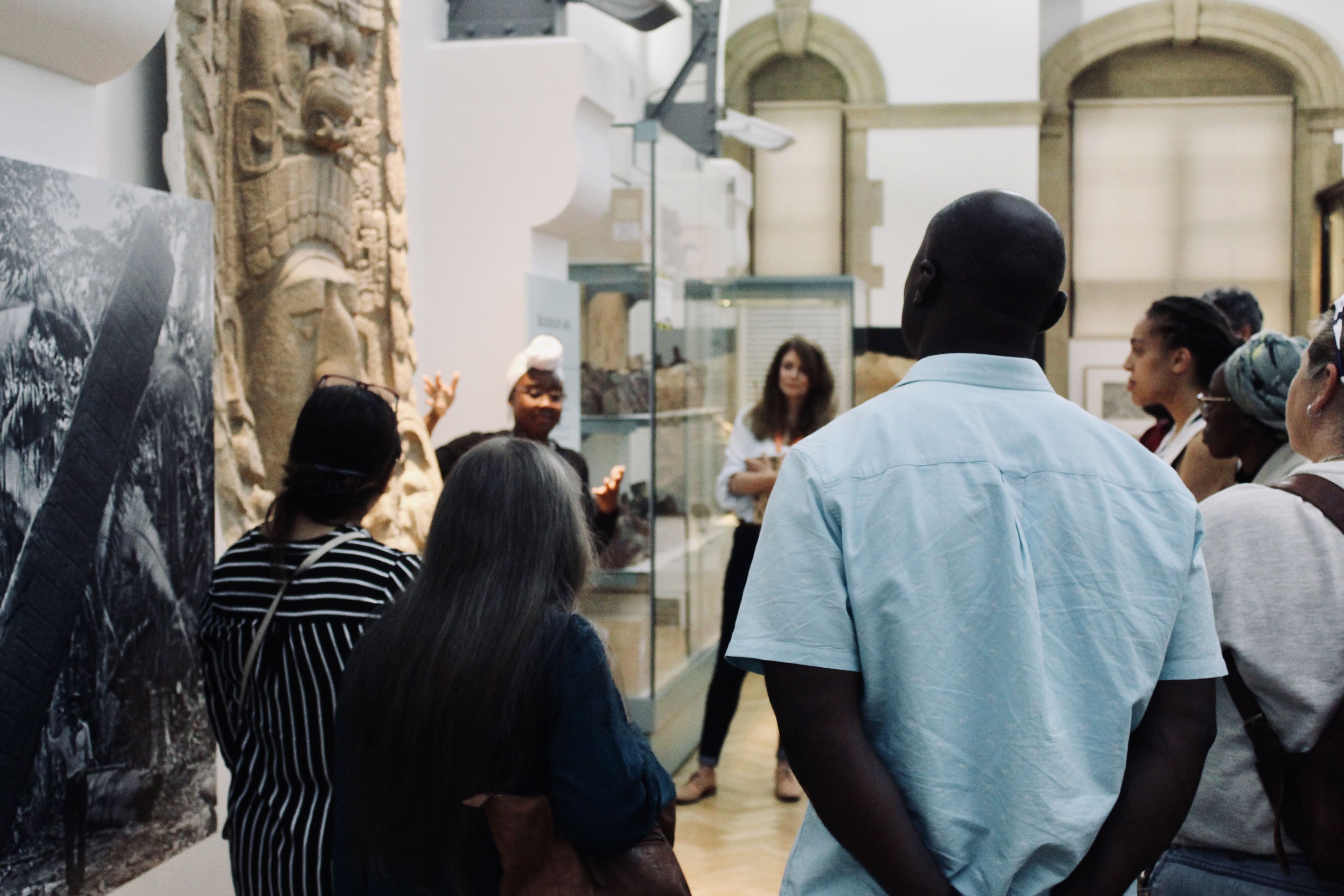 A group of people stand listening to a black woman wearing a white headscarf who is speaking and gesturing towards a stone sculpture at the Museum of Archaeology and Anthropology.