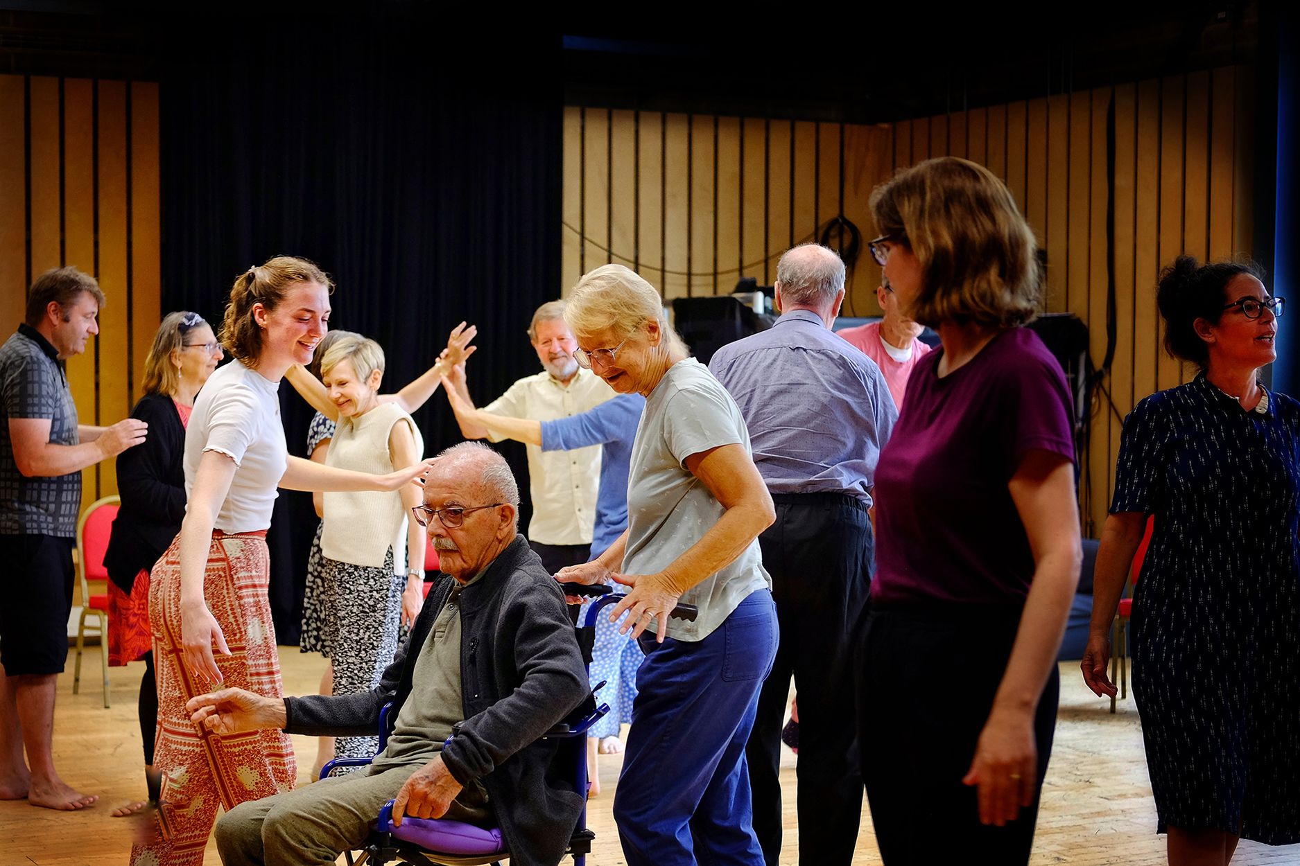 A group of people, some standing, one in a wheelchair, dance around in a room at Cambridge Junction.