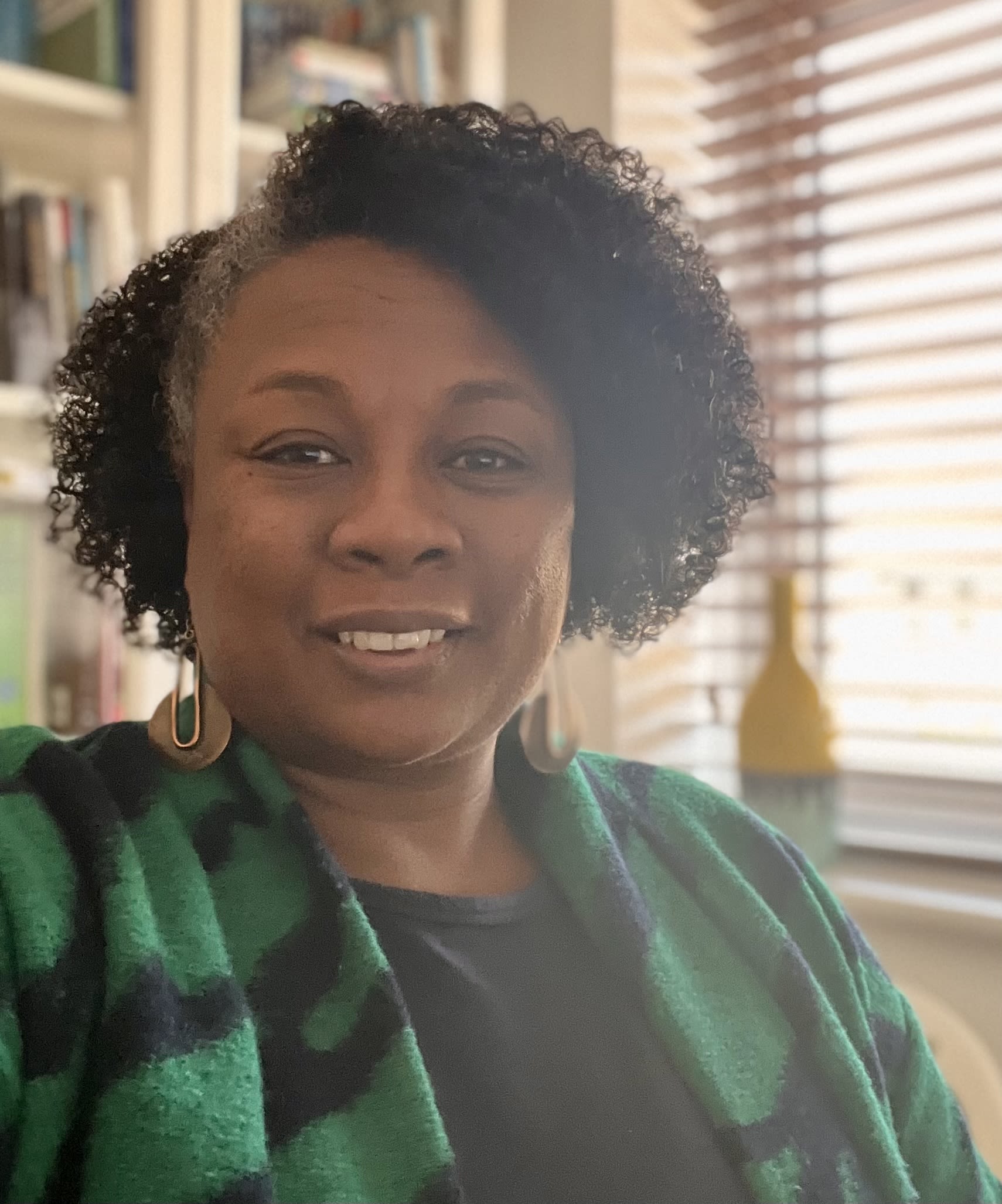 Akua, a black woman wearing a green cardigan smiles in a room with light coming through open blinds and a bookshelf behind.