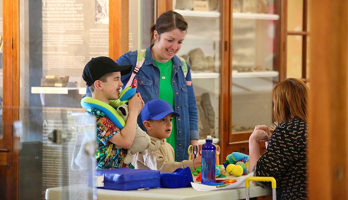 A smiling woman and two young boys stand in front of a table being welcomed by a member of UCM staff to the Sedgwick Museum. Wooden and glass display cases are visible in the background.