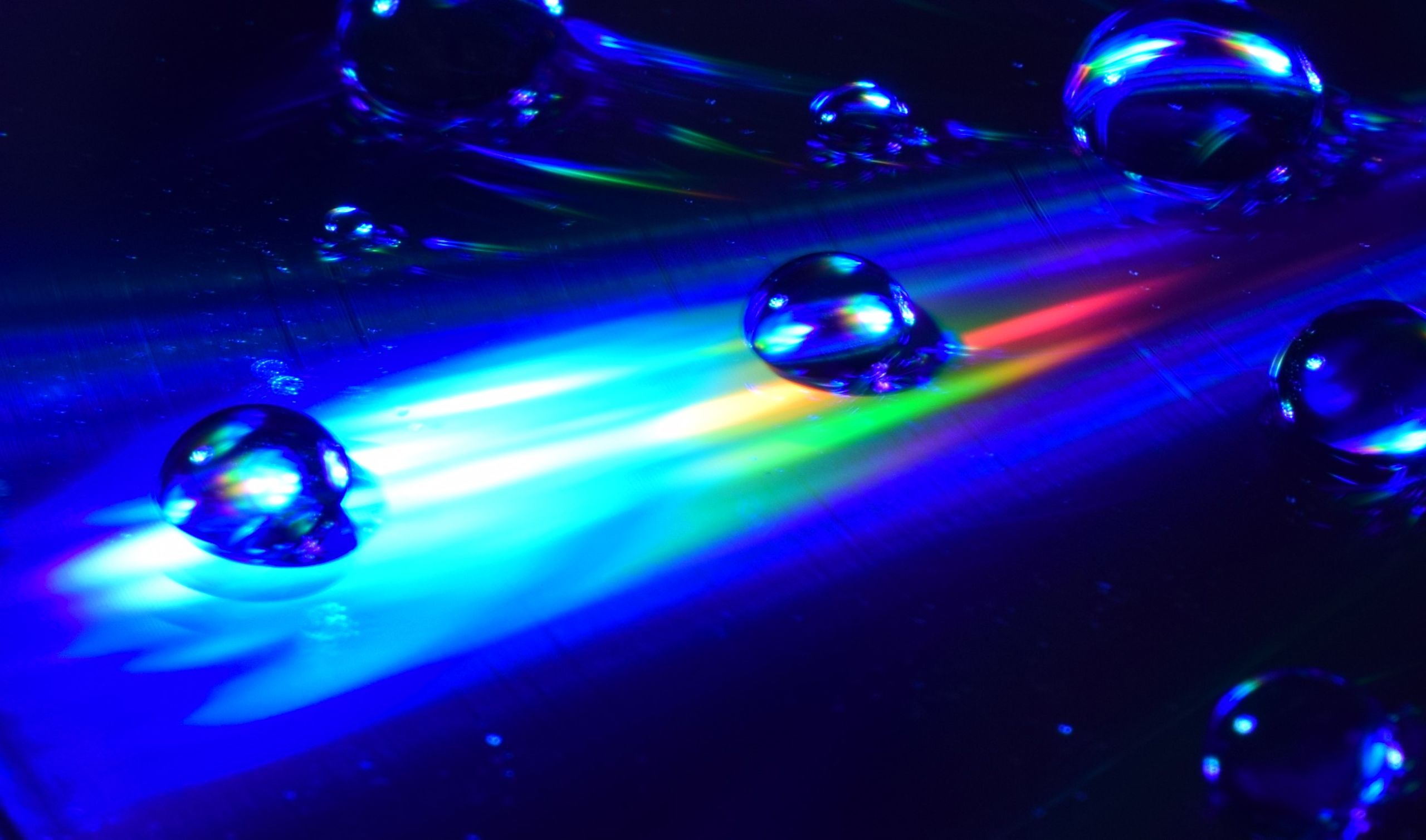 Water droplets lit by blue light, on a blue surface with steaks of light blue, yellow, green and red.